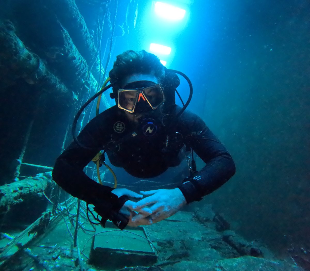 A scuba diver underwater in a deep blue environment, wearing a black wetsuit, mask, and breathing apparatus, with the diver holding their hands together.