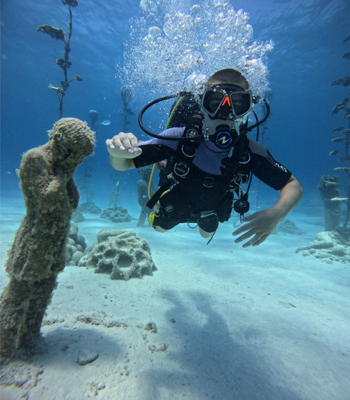 A diver underwater with a scuba mask and regulator, flexing his right arm, near a sandy ocean floor with coral and sea plants in the background.