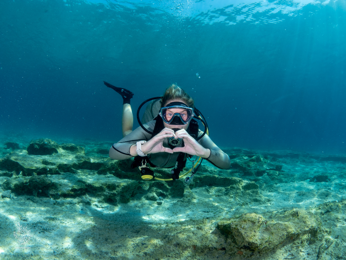 A woman scuba diving underwater, wearing a diving mask and wetsuit, holding a camera and making a heart shape with her hands, with rocks and sandy ocean floor below.