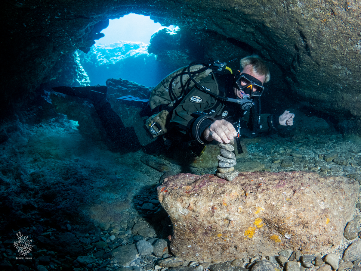 A scuba diver underwater in a cave, wearing a diving mask and suit, balancing a small rock stack on a large rock surface.
