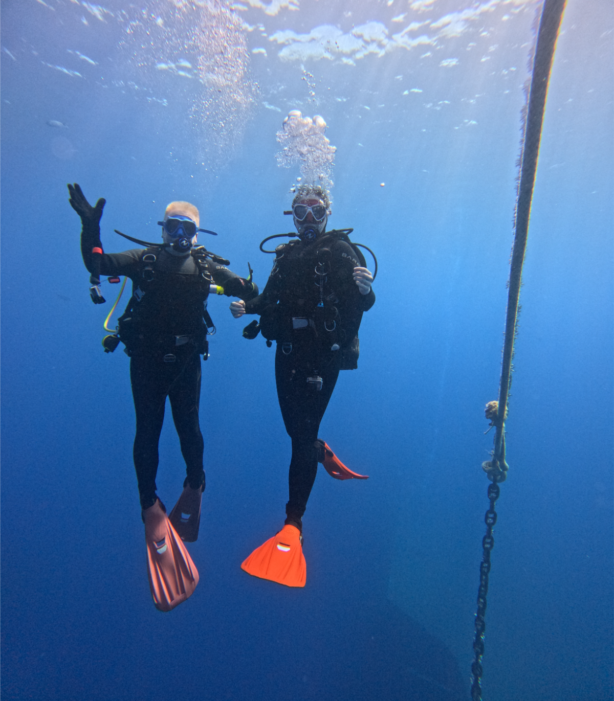 Two scuba divers in wetsuits and snorkel gear underwater, posing for a photo with a chain hanging beside them.