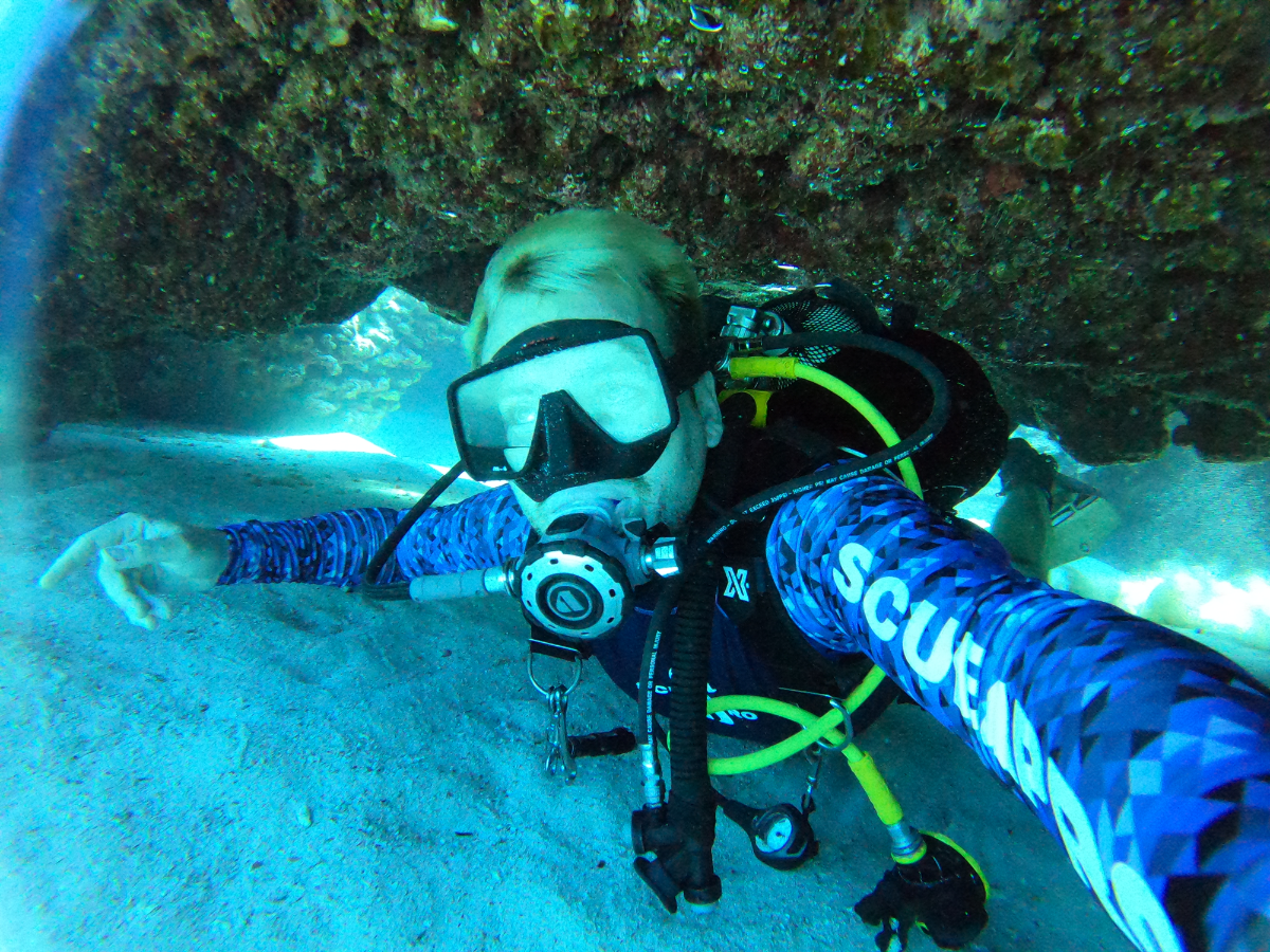 Scuba diver underwater taking a selfie with a rocky ceiling above and sandy floor below, wearing a mask, regulator, and a blue wetsuit with 'SCUBA' written on the sleeve.