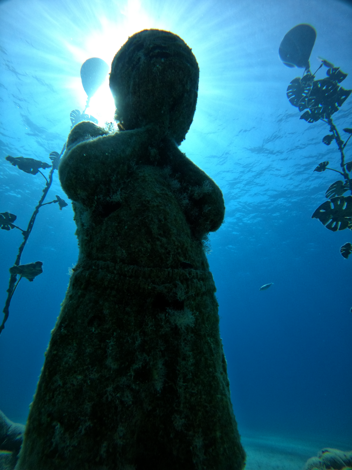 A submerged statue of a person holding balloons, covered in marine growth, with sunlight shining above and fish swimming nearby.