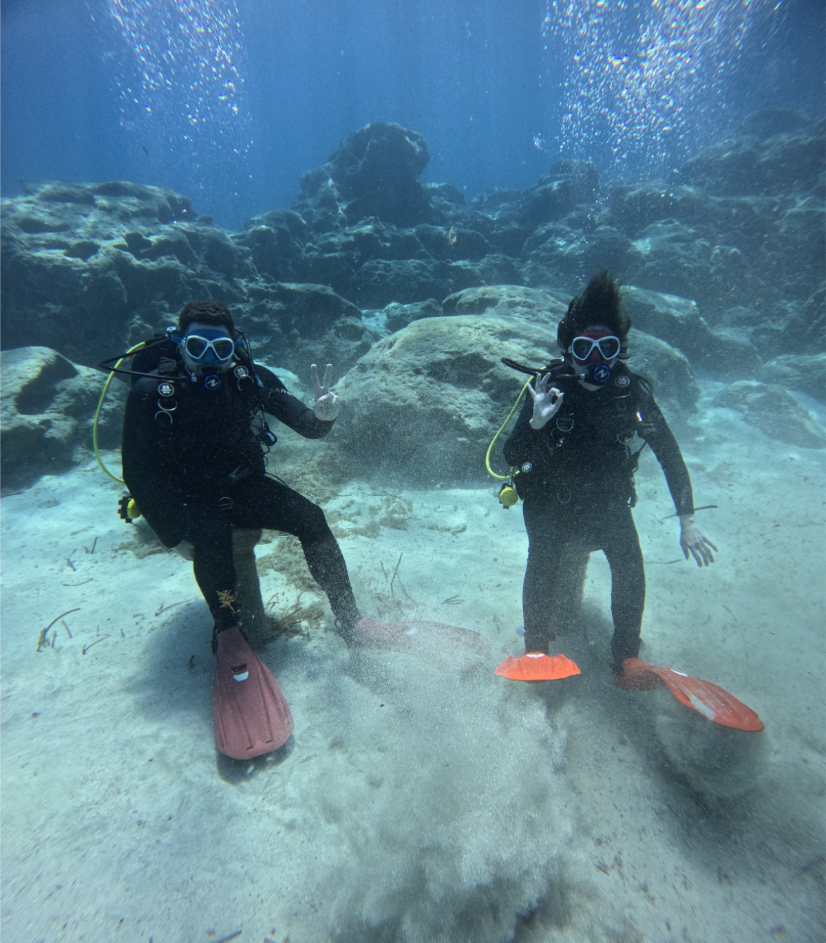 Two divers underwater on a sandy seabed with rocks in the background, making hand signals and wearing scuba gear and orange fins.