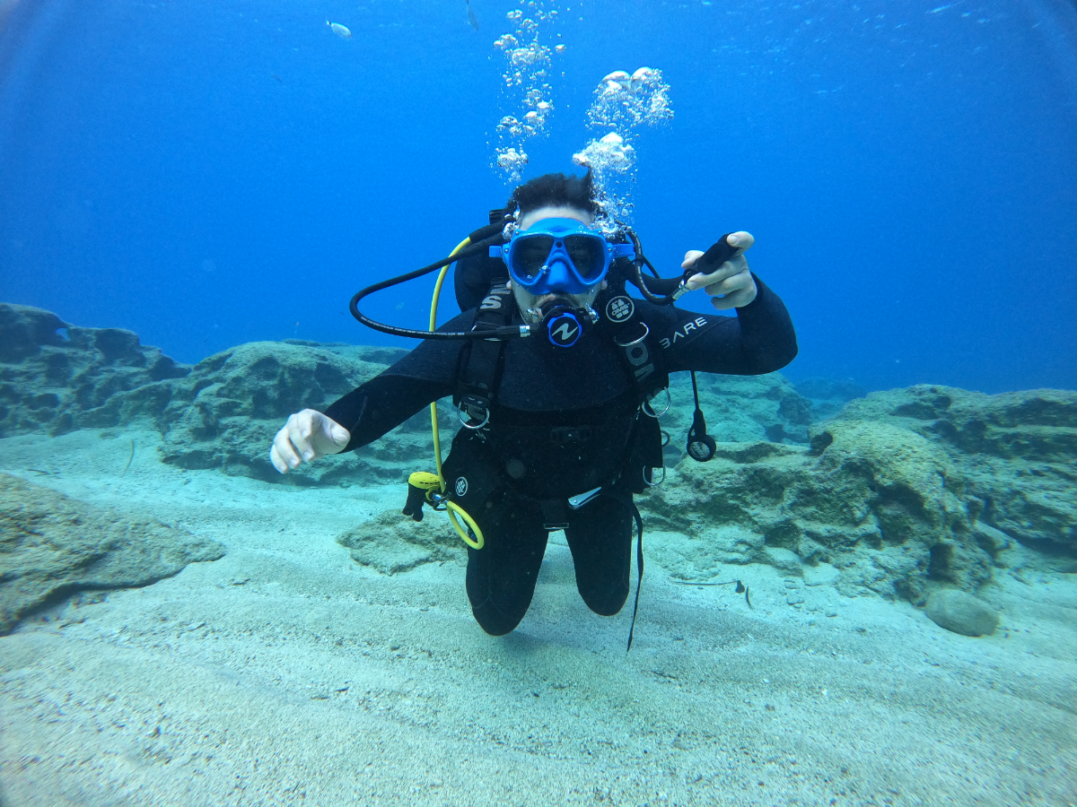 Person scuba diving underwater, wearing a black wetsuit, blue mask, and scuba gear, pointing at the camera with a rocky seabed in the background.