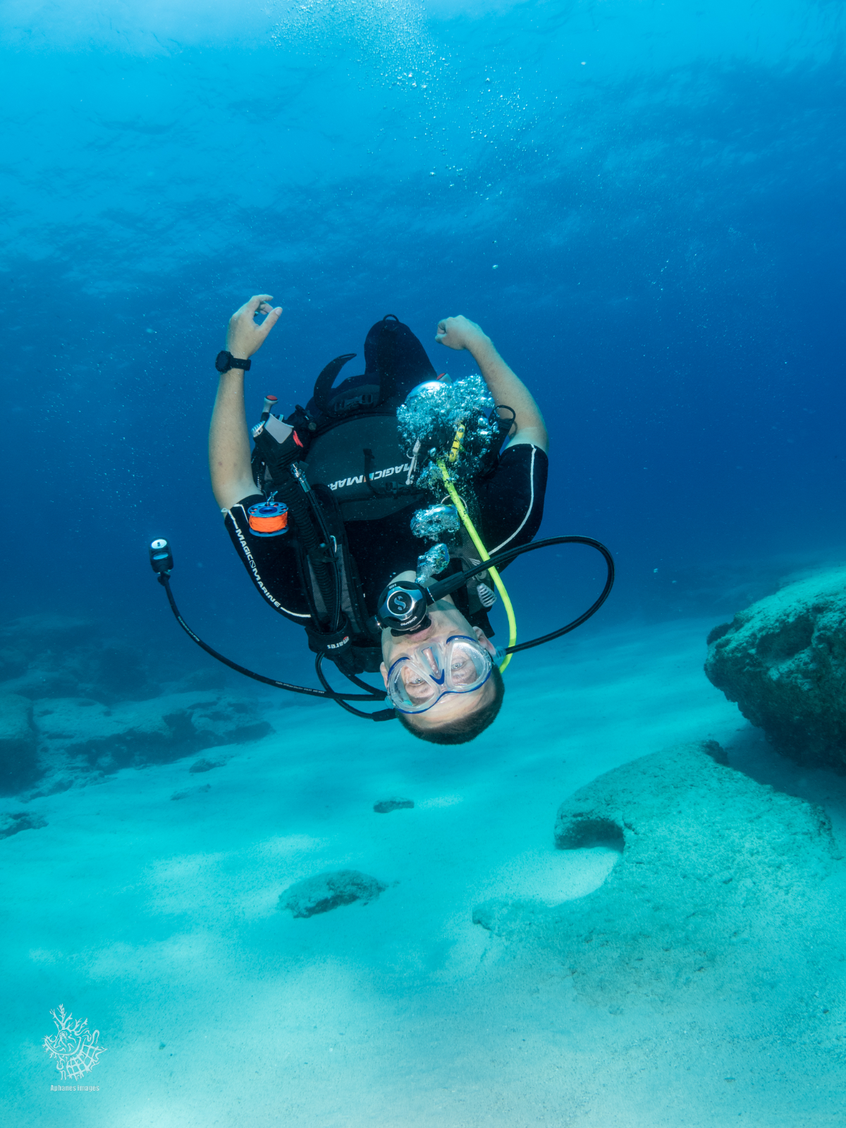 A scuba diver underwater wearing a mask and breathing apparatus, near sandy ocean floor with rocks.