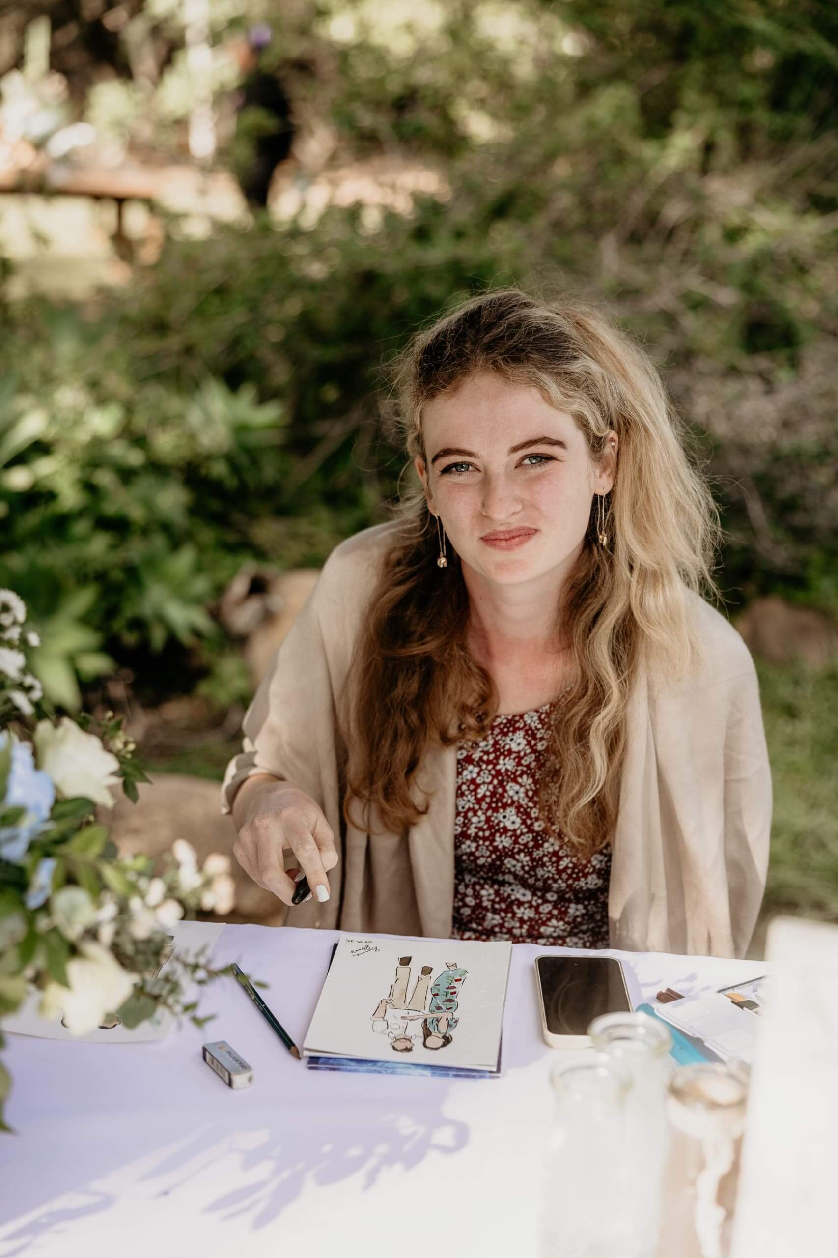A woman with wavy blonde hair, wearing a patterned top and beige blazer, sitting at a table outdoors with art supplies, a smartphone, and a floral centerpiece, looking at the camera with a neutral expression.