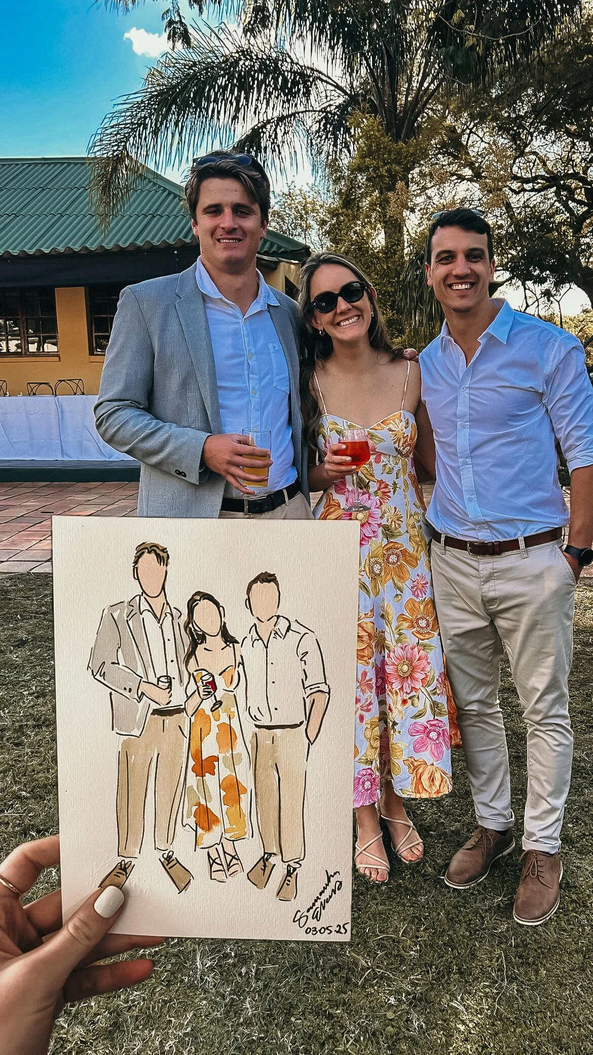 Group of three people standing outdoors at an event, with a palm tree and building in the background. They are dressed in semi-formal attire, holding drinks, smiling for the photo. A hand is holding a sketch art of the same three people in the foregr