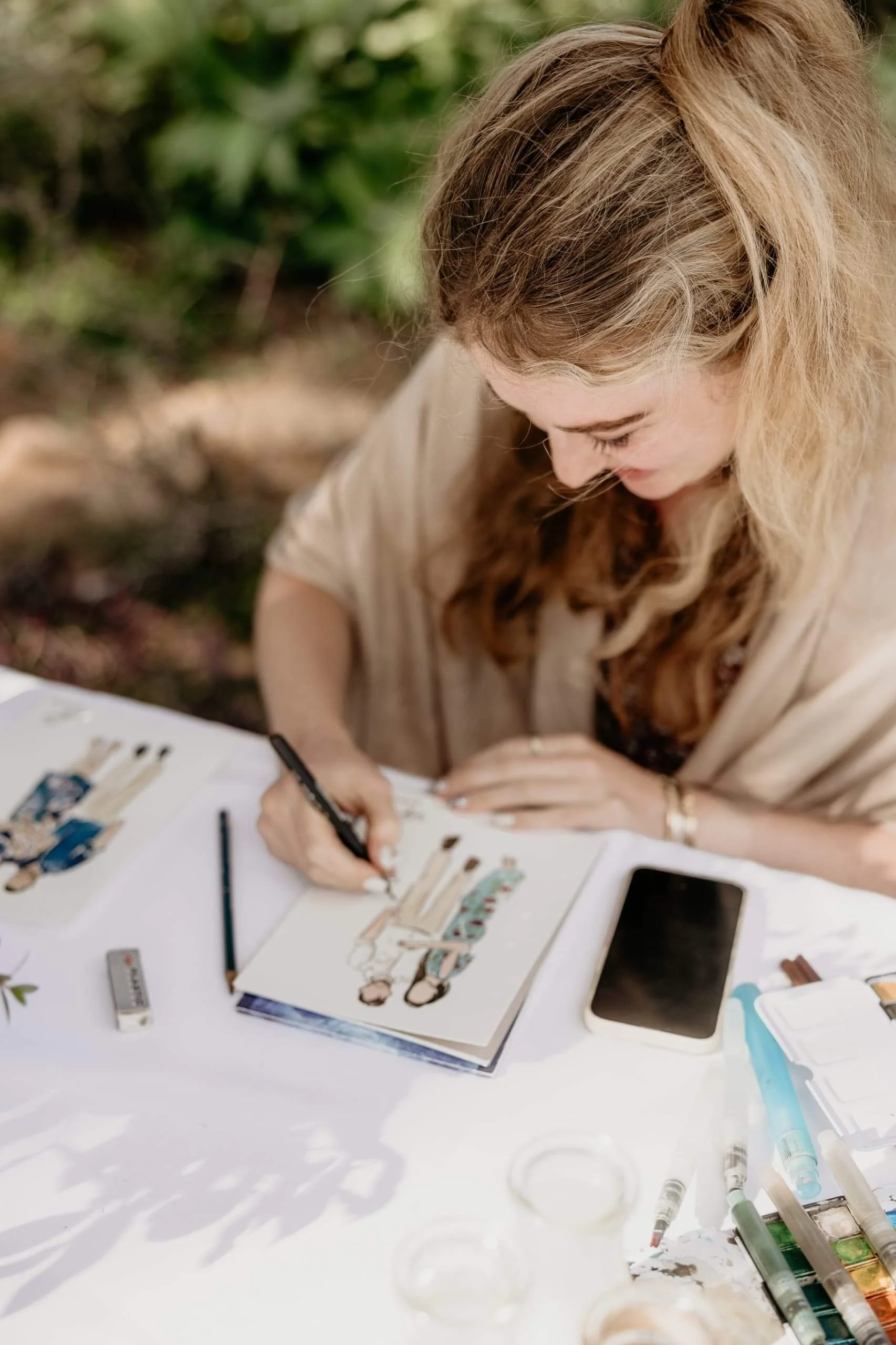 Woman drawing fashion sketches at an outdoor table, like a live wedding portrait artist Zimbabwe, surrounded by art supplies and greenery.