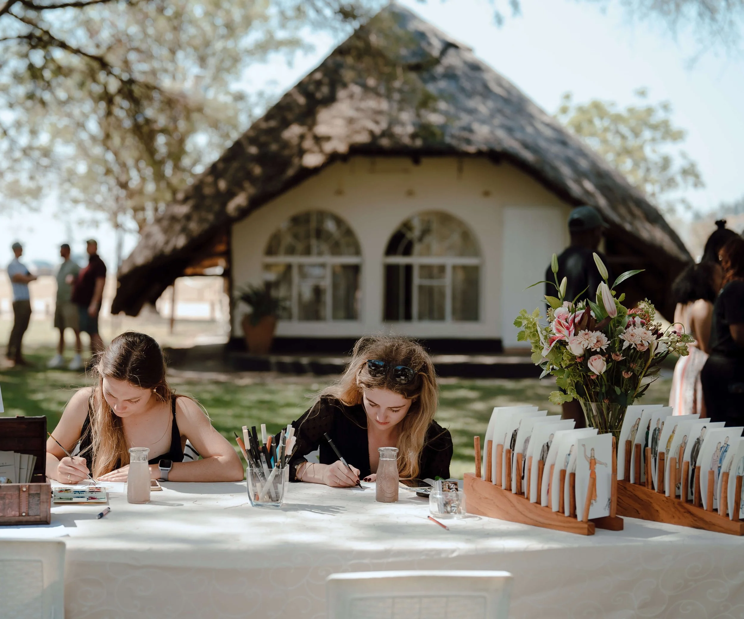 Two women sitting at a table outdoors, coloring with markers. The table has a large floral arrangement and a display of illustrated cards. A thatched roof house and people chatting are in the background.