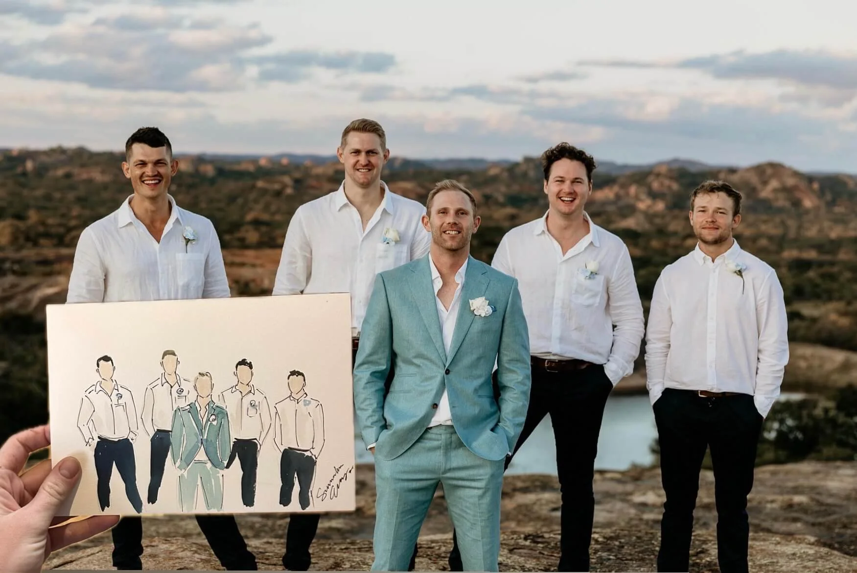 A group of five men dressed in white shirts, with one wearing a light blue suit, standing outdoors on a rocky landscape with a river and hills in the background, smiling at the camera. The scene appears to be during sunset or late afternoon.