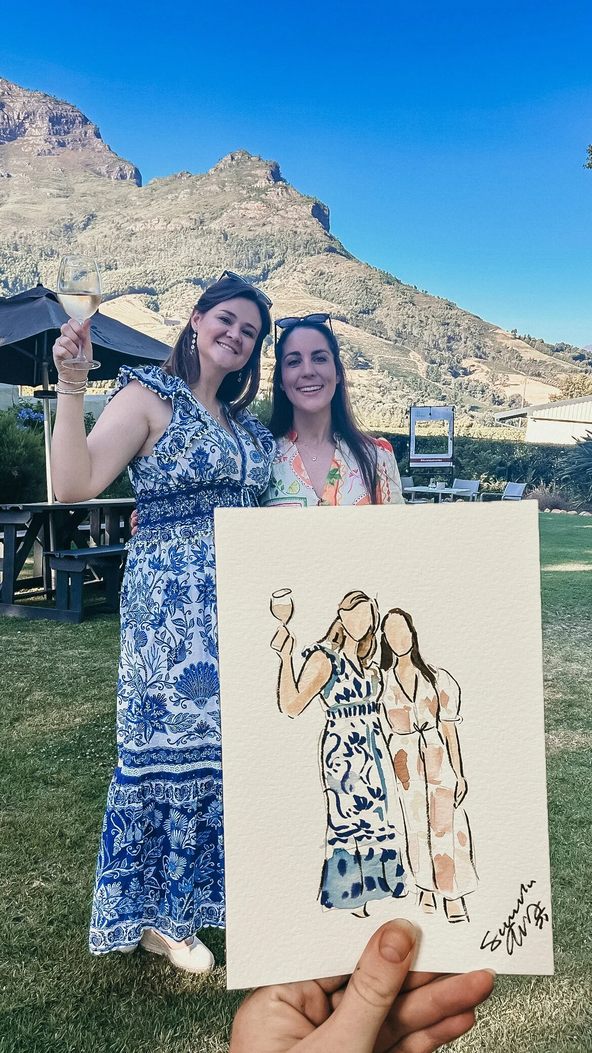 Two women standing outdoors with a mountain in the background, one holding a glass of champagne. An artists holds a watercolor sketch of the two women, at a destination wedding portrait Artist event