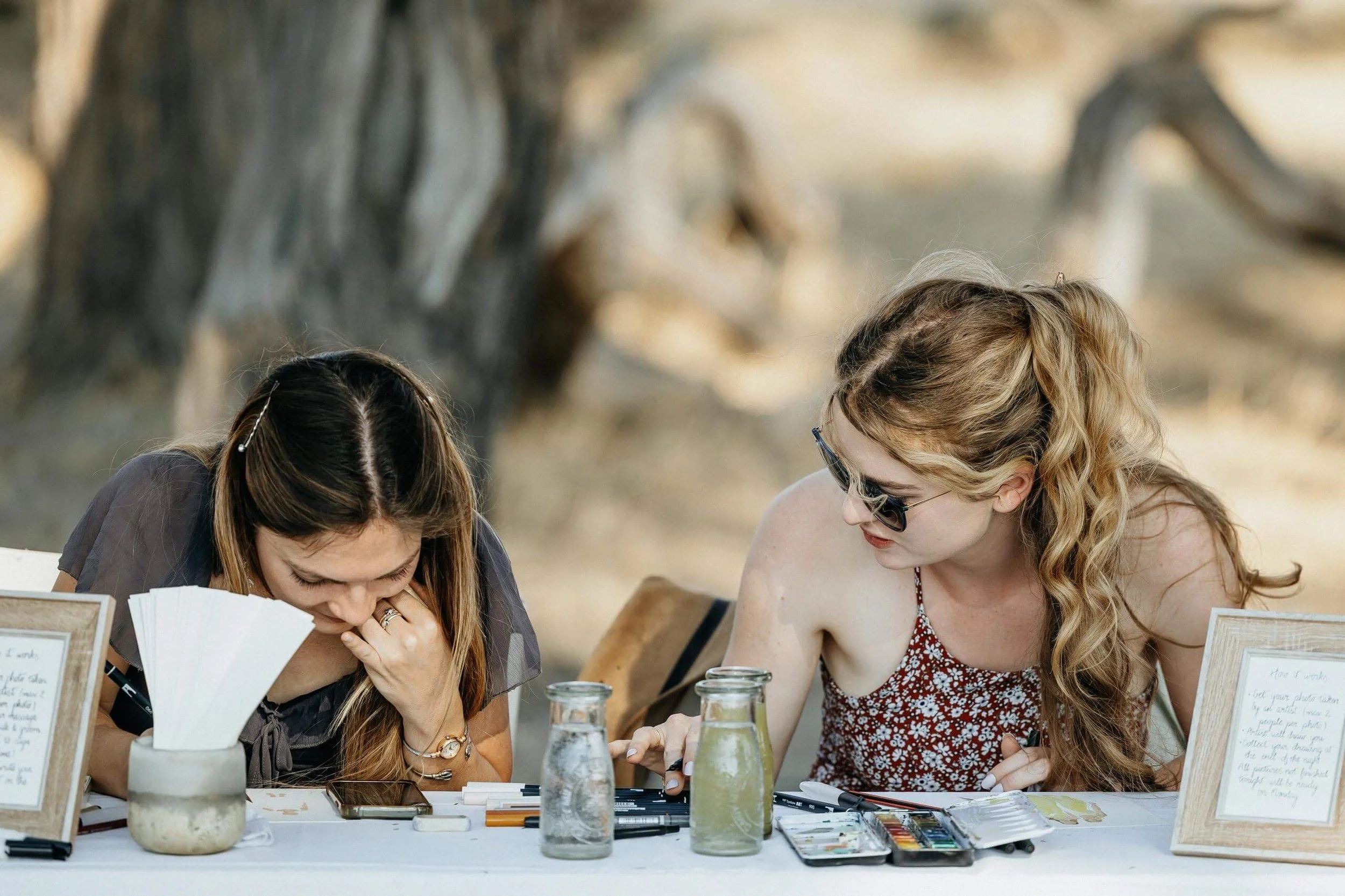 Two women sit at a table outdoors with art supplies, engaged in a creative activity, with trees in the background.