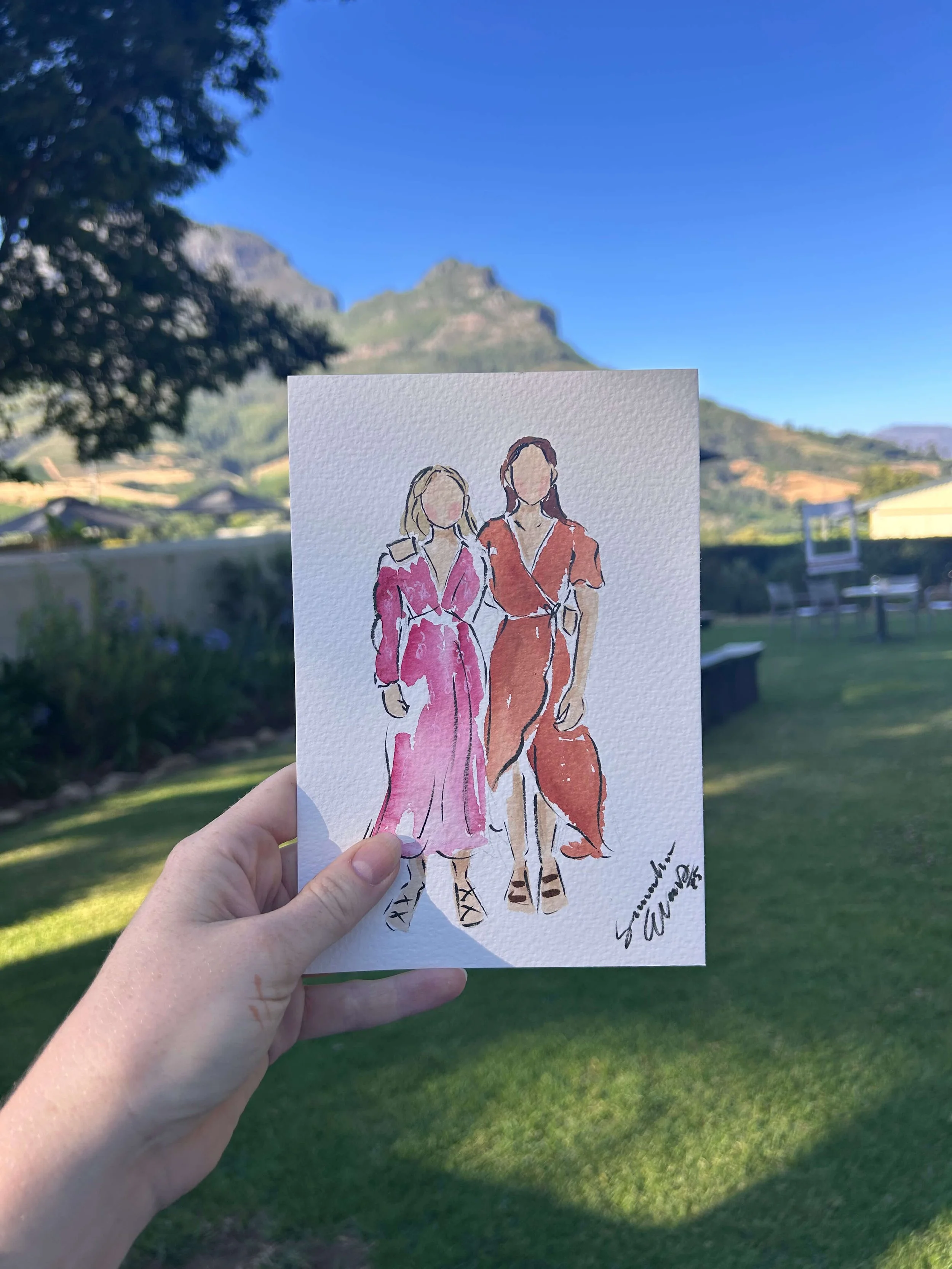Hand holding a watercolor illustration of two women in stylish dresses outdoors with a scenic mountain background.