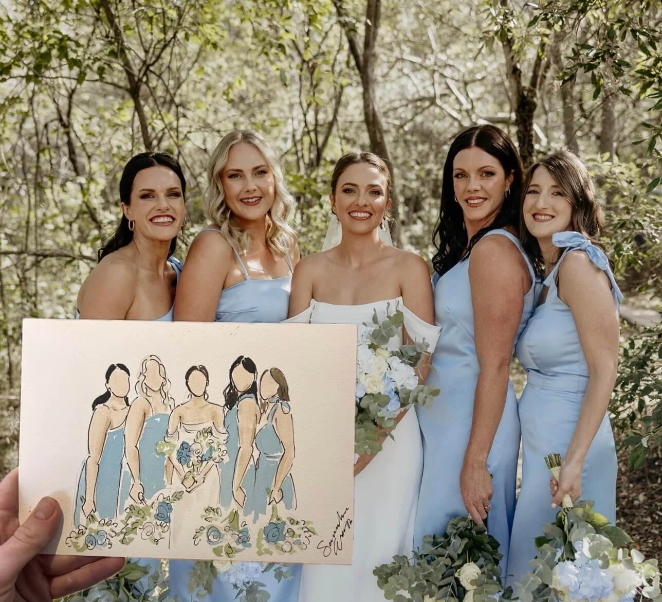 Live Event Illustrator for Weddings painting a group of five women dressed in light blue dresses standing outdoors among trees, with one woman in a white wedding dress holding a bouquet of white and blue flowers.