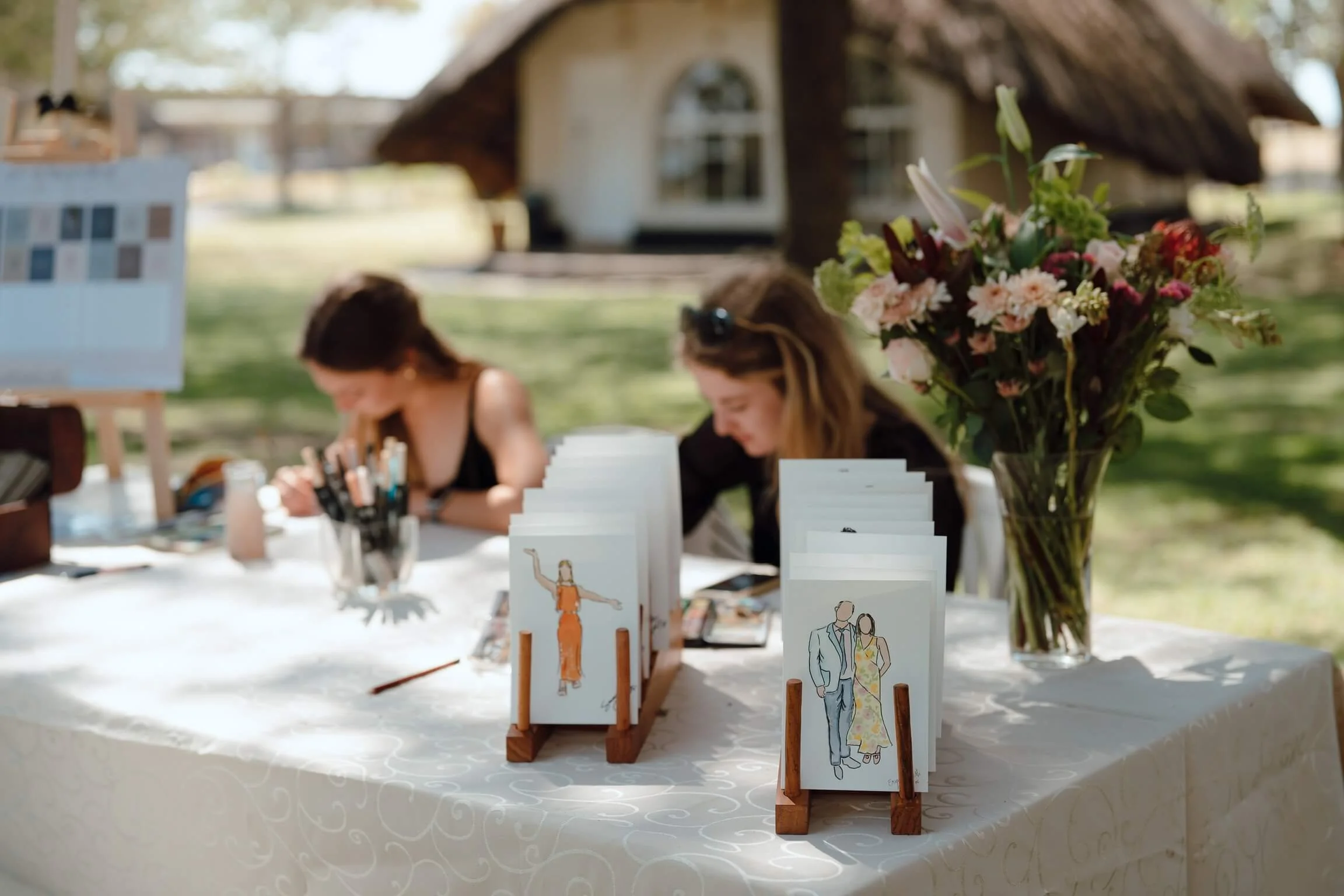 Table with wedding or event invitations, flower arrangement, and art supplies outdoors with blurred house in background.