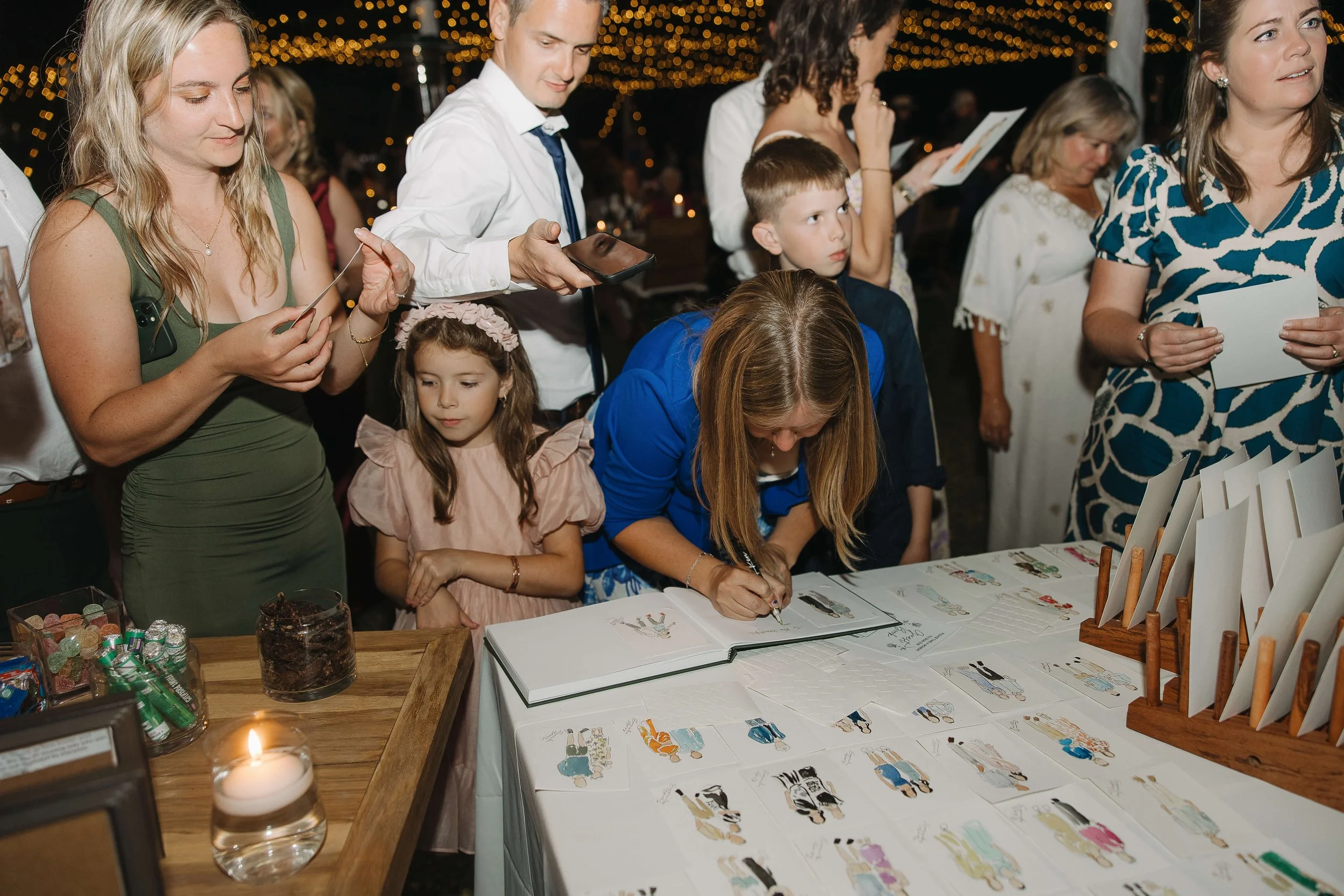 A woman looking at live custom portrait painting at a wedding reception while surrounded by guests, including children, under string lights at night.