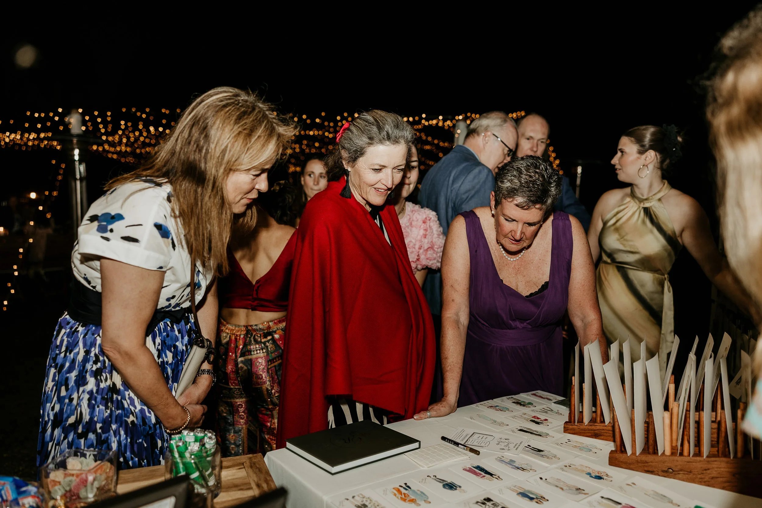 Group of people at a nighttime event looking at a table with artwork and photographs.