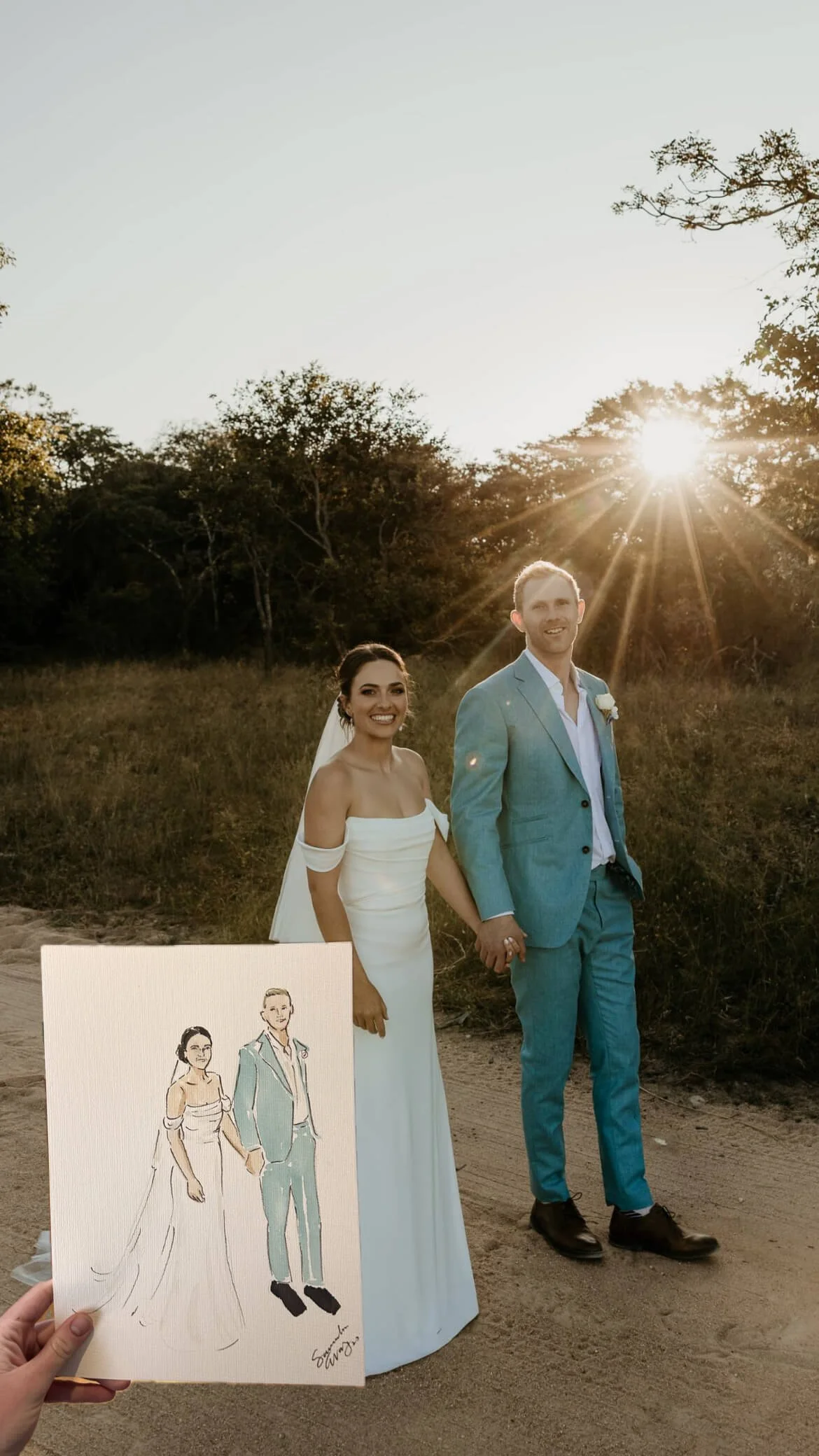 A bride and groom holding hands in a field during sunset, with a drawing of themselves held by a person in the foreground.