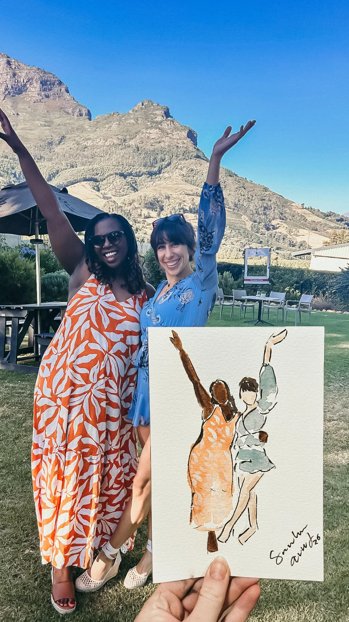 Two women standing outdoors in front of mountainous landscape, one in an orange and white patterned dress, the other in a blue floral dress, smiling and raising their arms. Someone is holding a watercolor painting of the same scene, labeled "Save the