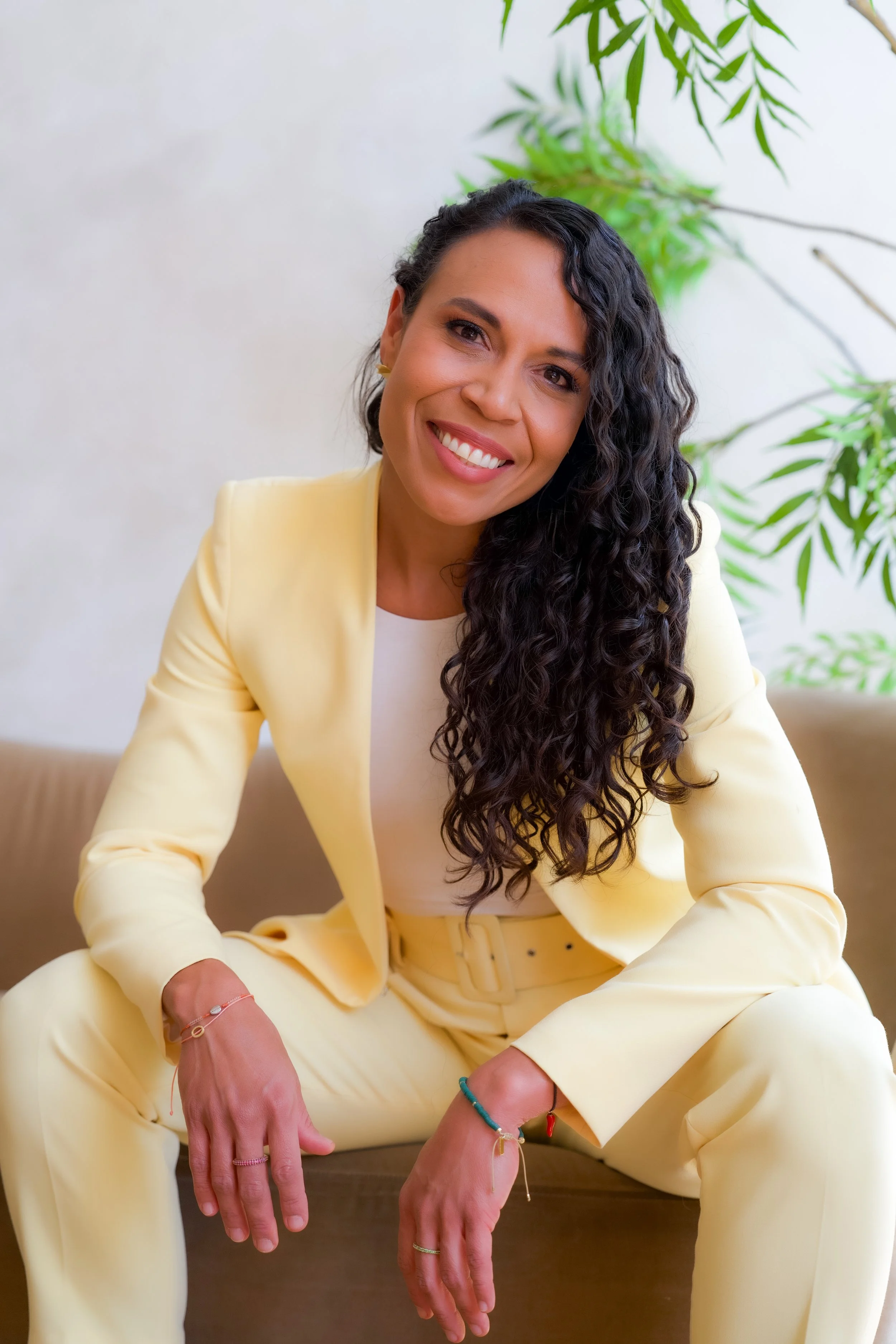 A woman with long, curly black hair, smiling and sitting on the floor, wearing a light yellow suit and sitting in front of a green leafy plant, with a white background.