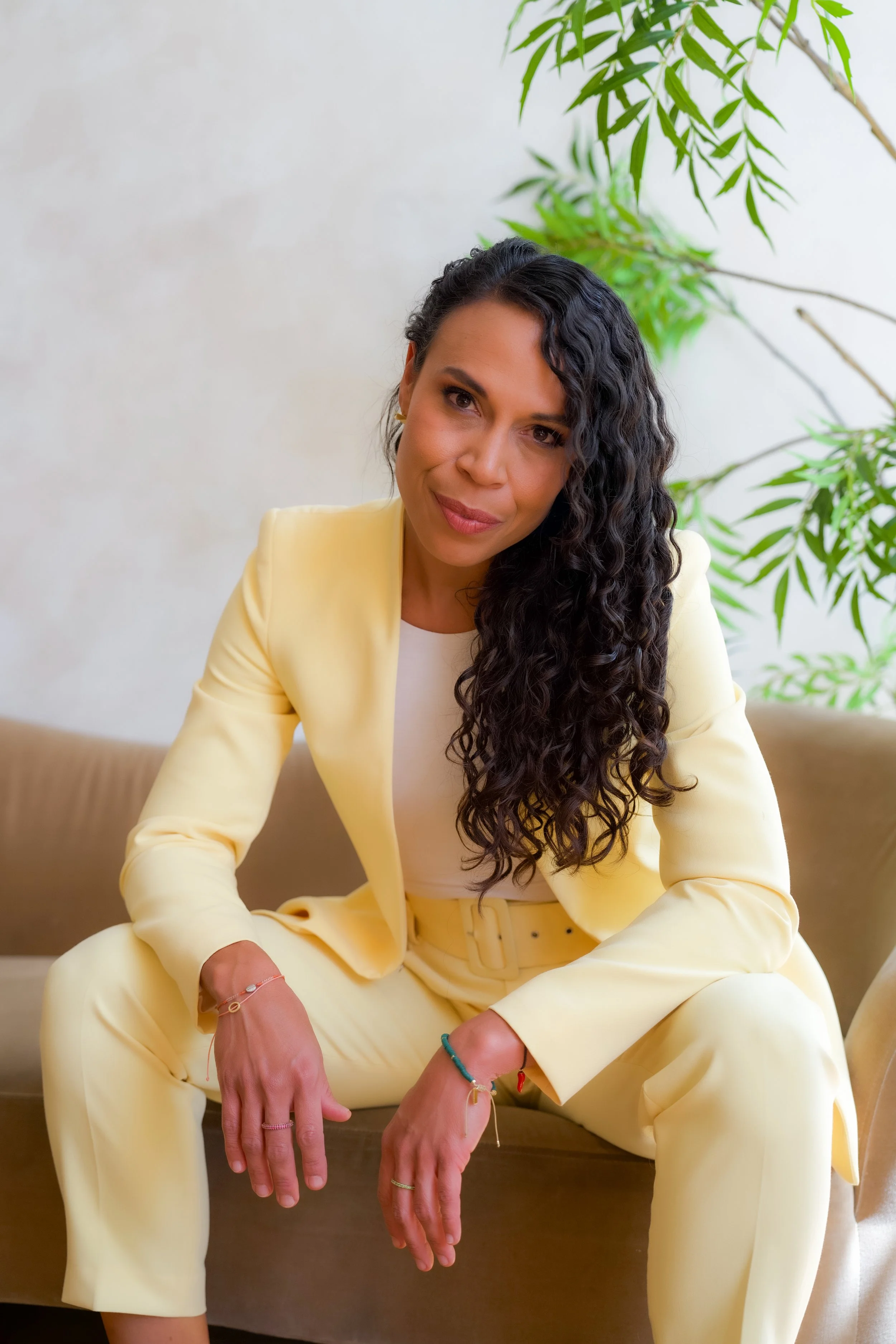 Woman with dark curly hair wearing a yellow suit sitting on a beige couch with a green plant in the background.