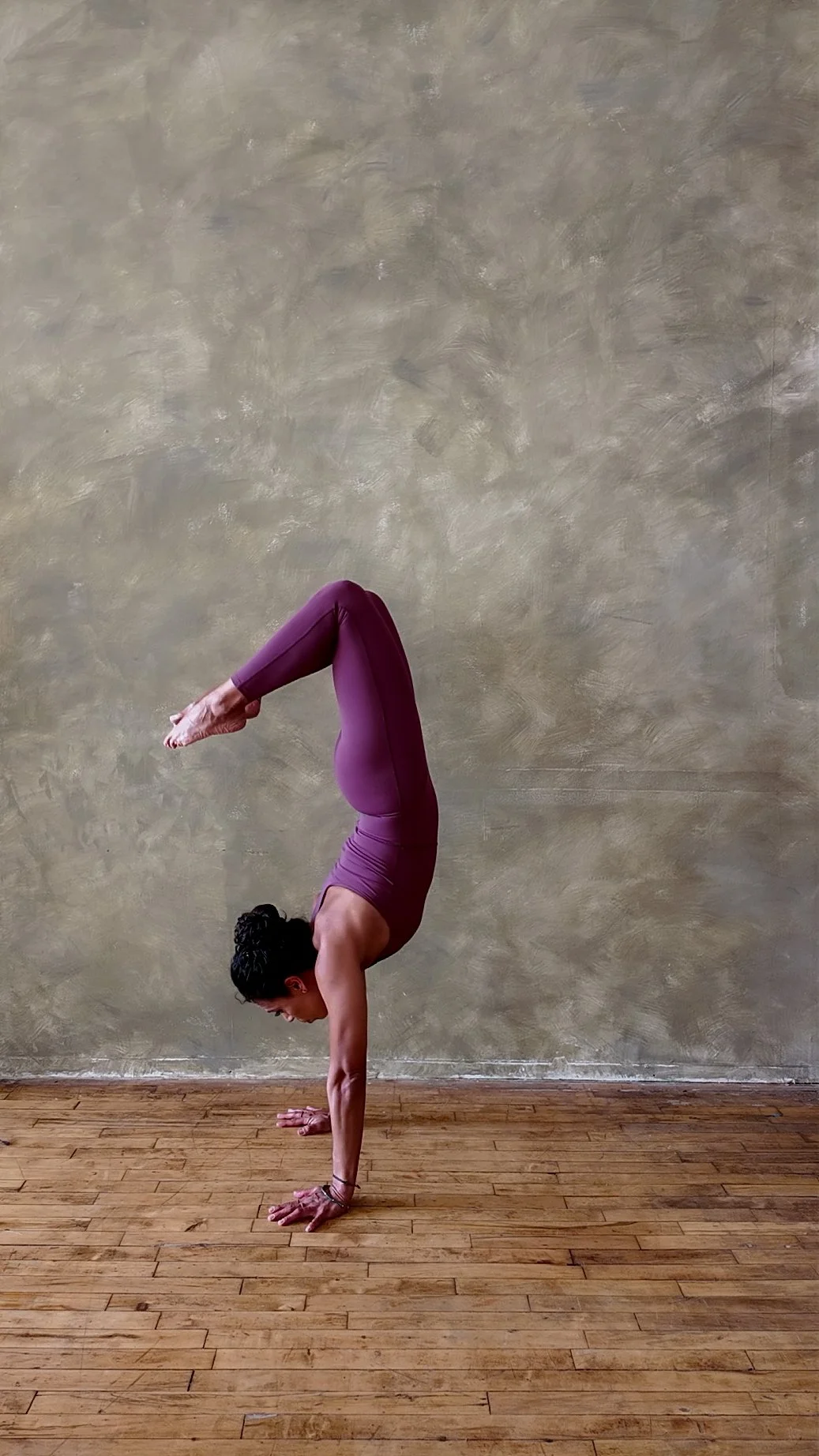 A woman in purple athletic wear practicing a handstand yoga pose indoors on wooden flooring against a textured beige wall.