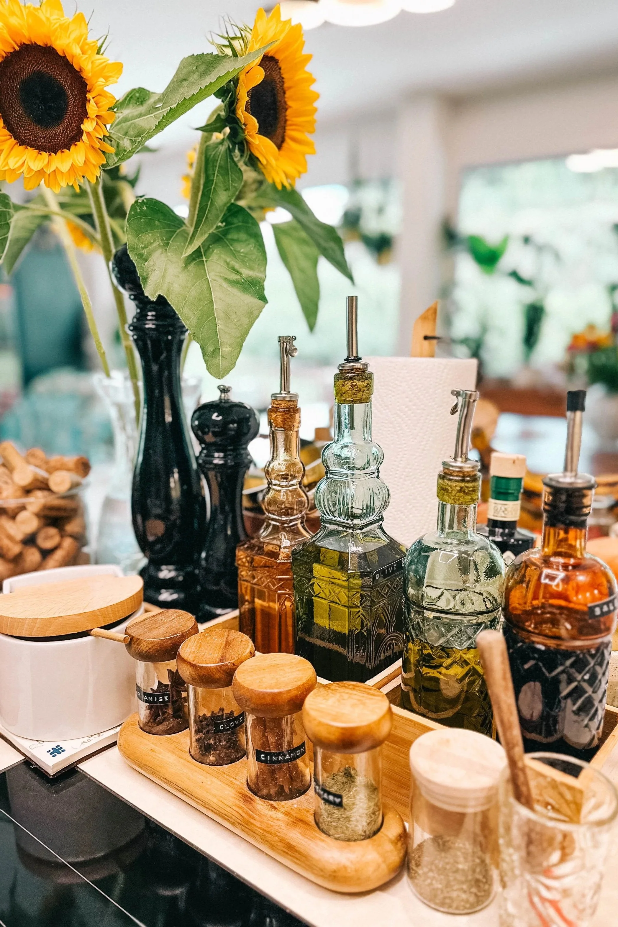 A collection of decorative glass bottles with wooden stoppers, small spice jars labeled with herbs and spices, a black vase with sunflowers, and various kitchen items on a countertop.