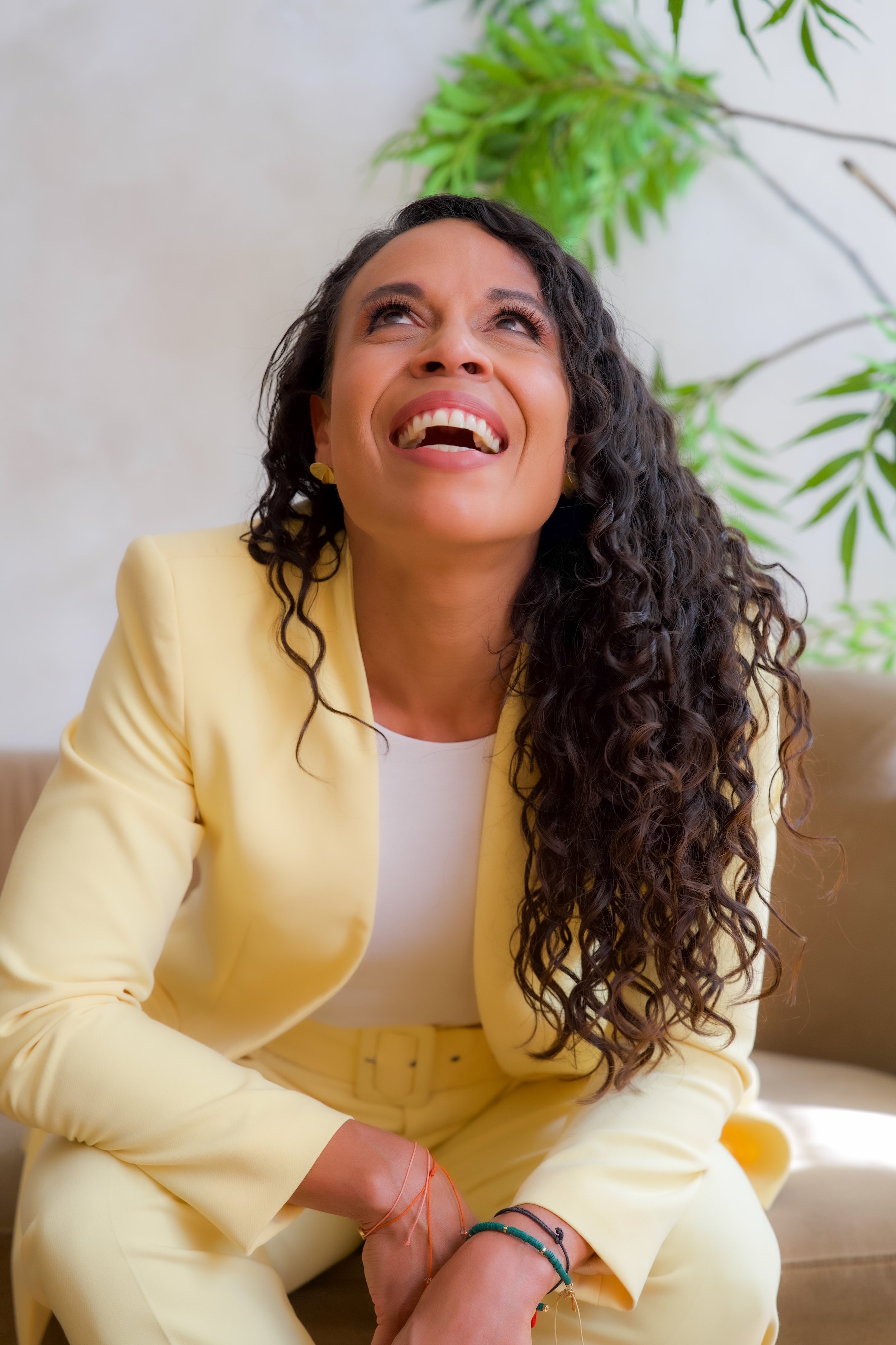 A woman with long curly hair, smiling and looking upward, wearing a yellow suit with a white top, sitting in front of a light-colored wall with green plants in the background.