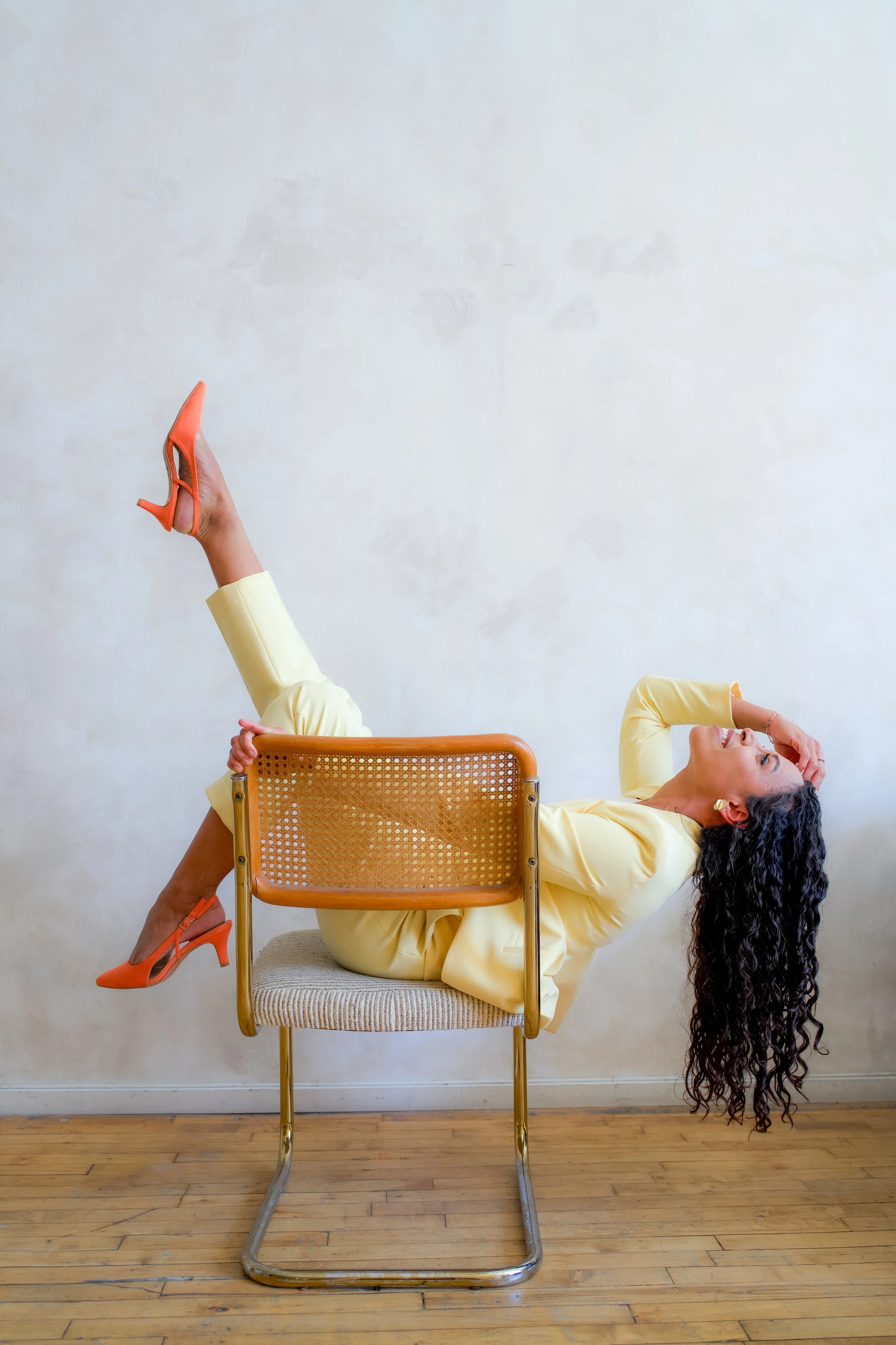 A woman wearing an oversized yellow blazer and matching pants is lying on her back on a wooden floor. Her head is tilted backward, and she has long dark curly hair cascading down. Her left leg is bent with the foot resting on the seat of a modern chair, and her right leg is raised in the air, wearing an orange high-heeled shoe. She appears to be smiling or laughing.