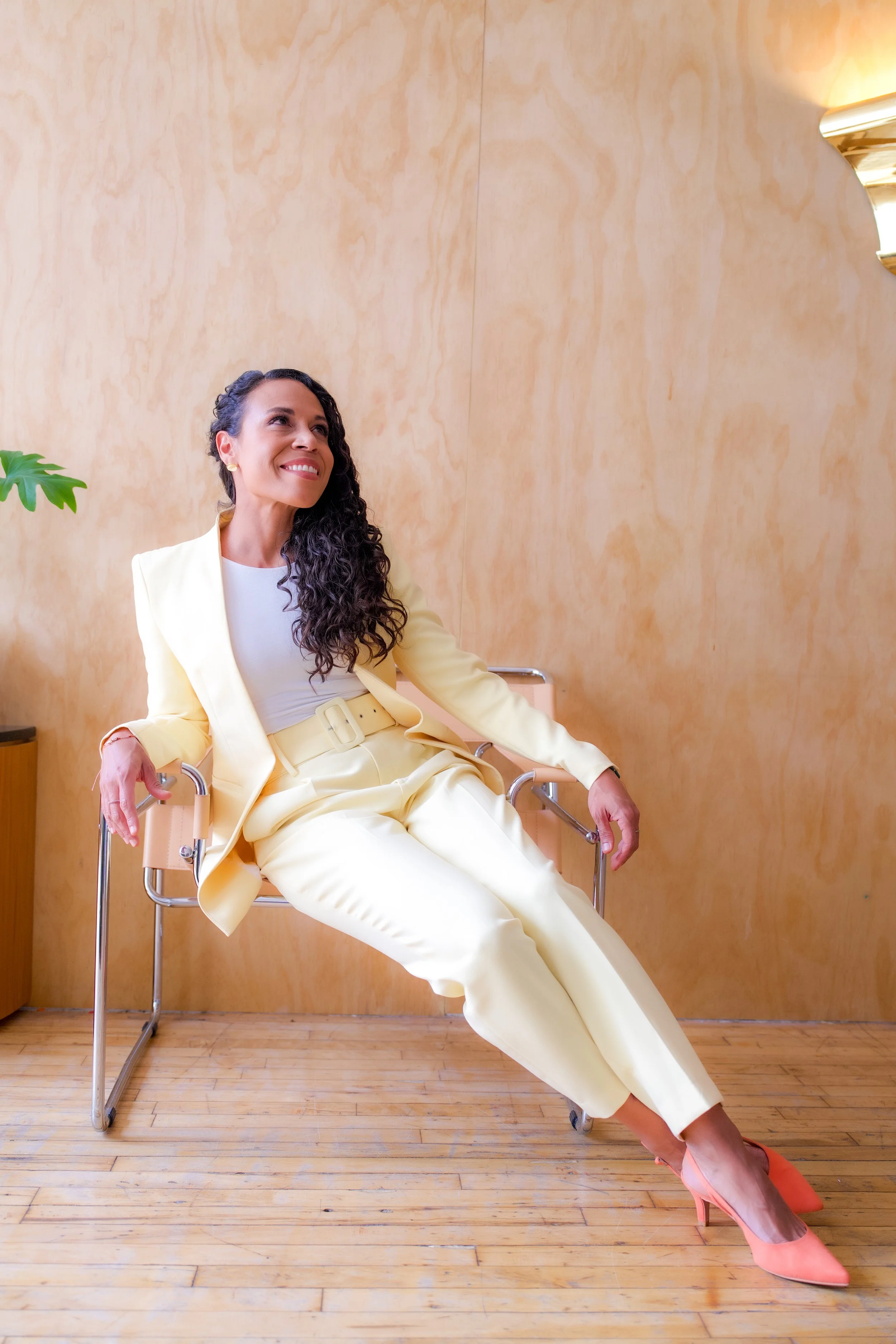 A woman in a yellow pantsuit sitting on a chair with her legs crossed, smiling and looking off to the side, in a room with wooden flooring and a beige wall, wearing pink high heel shoes.