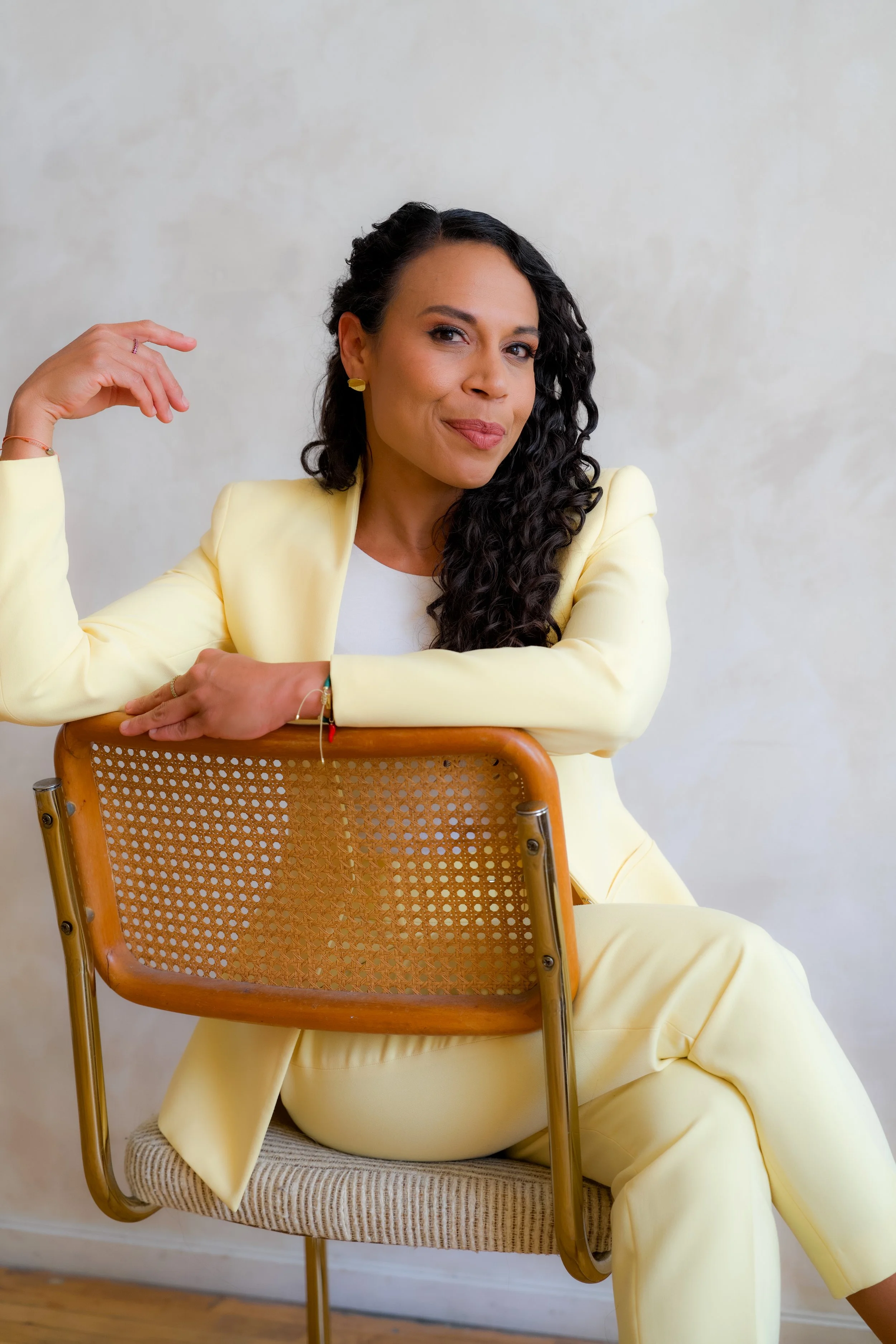 A woman with long, curly black hair is sitting on a chair, wearing a yellow suit and a white top. She is leaning on the back of a wooden chair with a woven cane backrest, resting one arm on it and smiling confidently.