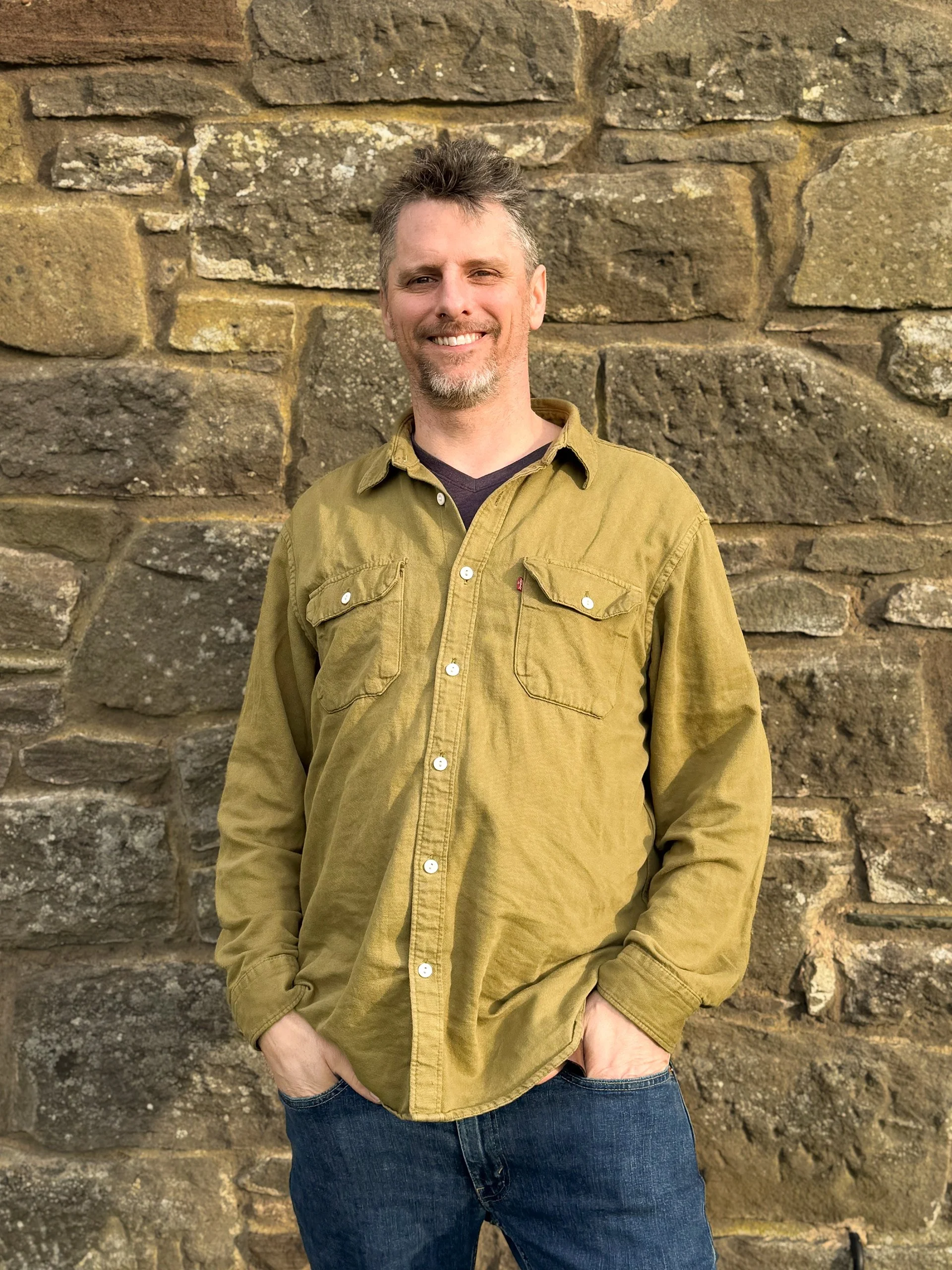 A man with short, spiky hair and a beard, wearing a mustard-colored button-up shirt with chest pockets, standing in front of a textured stone wall, smiling with hands in his pockets.