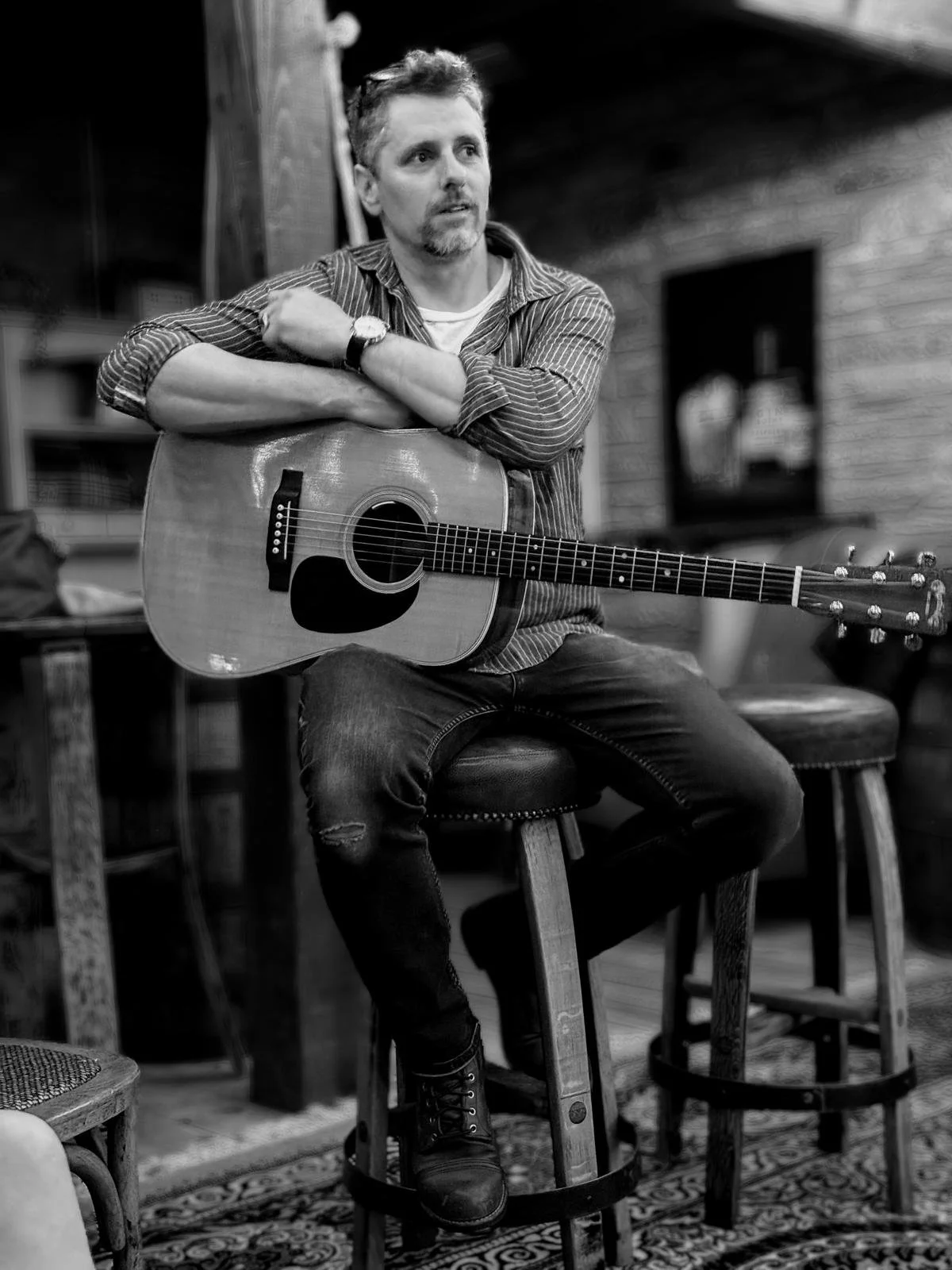 A man sitting on a stool with a guitar, looking thoughtful in a rustic indoor setting.
