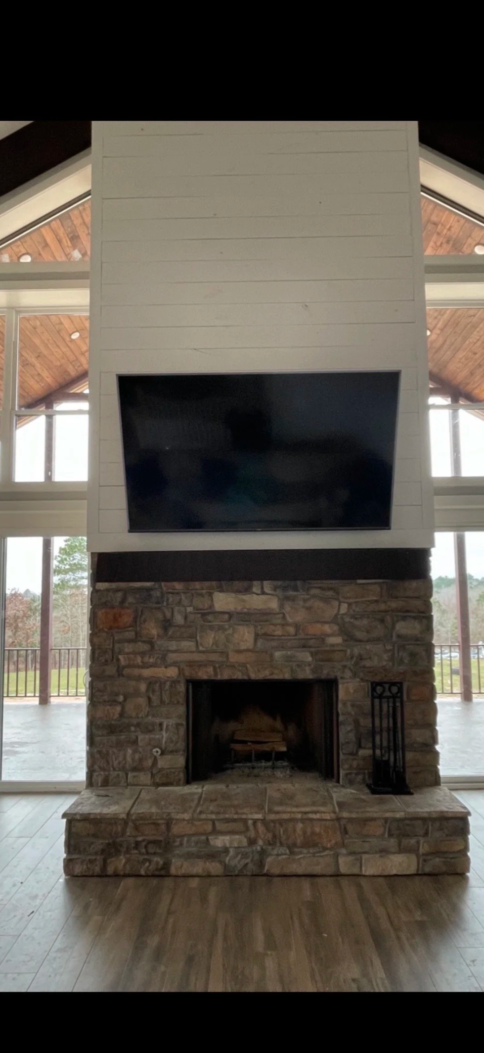 Living room fireplace with stone surround, a fireplace toolset on the right, and a large flat-screen TV above, with large windows and a wooden ceiling in the background.