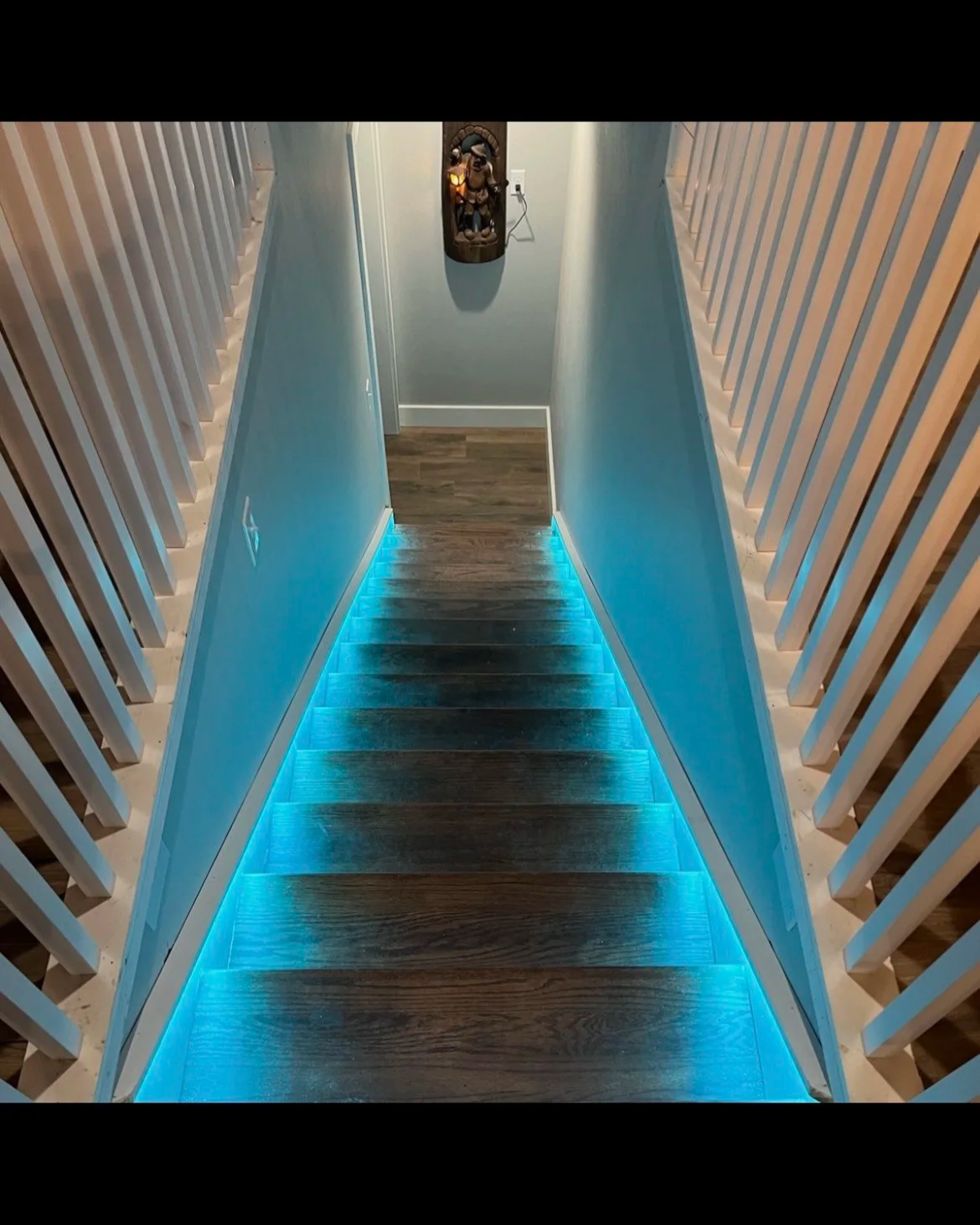 Downward view of a staircase with blue LED lighting under the steps, flanked by white railings on both sides, leading to a small landing with a wall-mounted decorative piece and an outlet.
