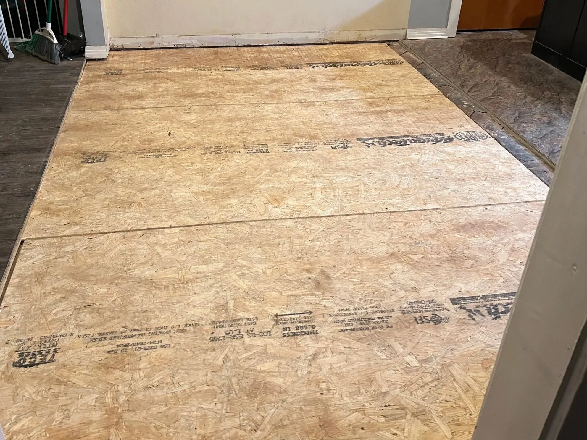 Plywood flooring in a room with a door and a transition to tile flooring.