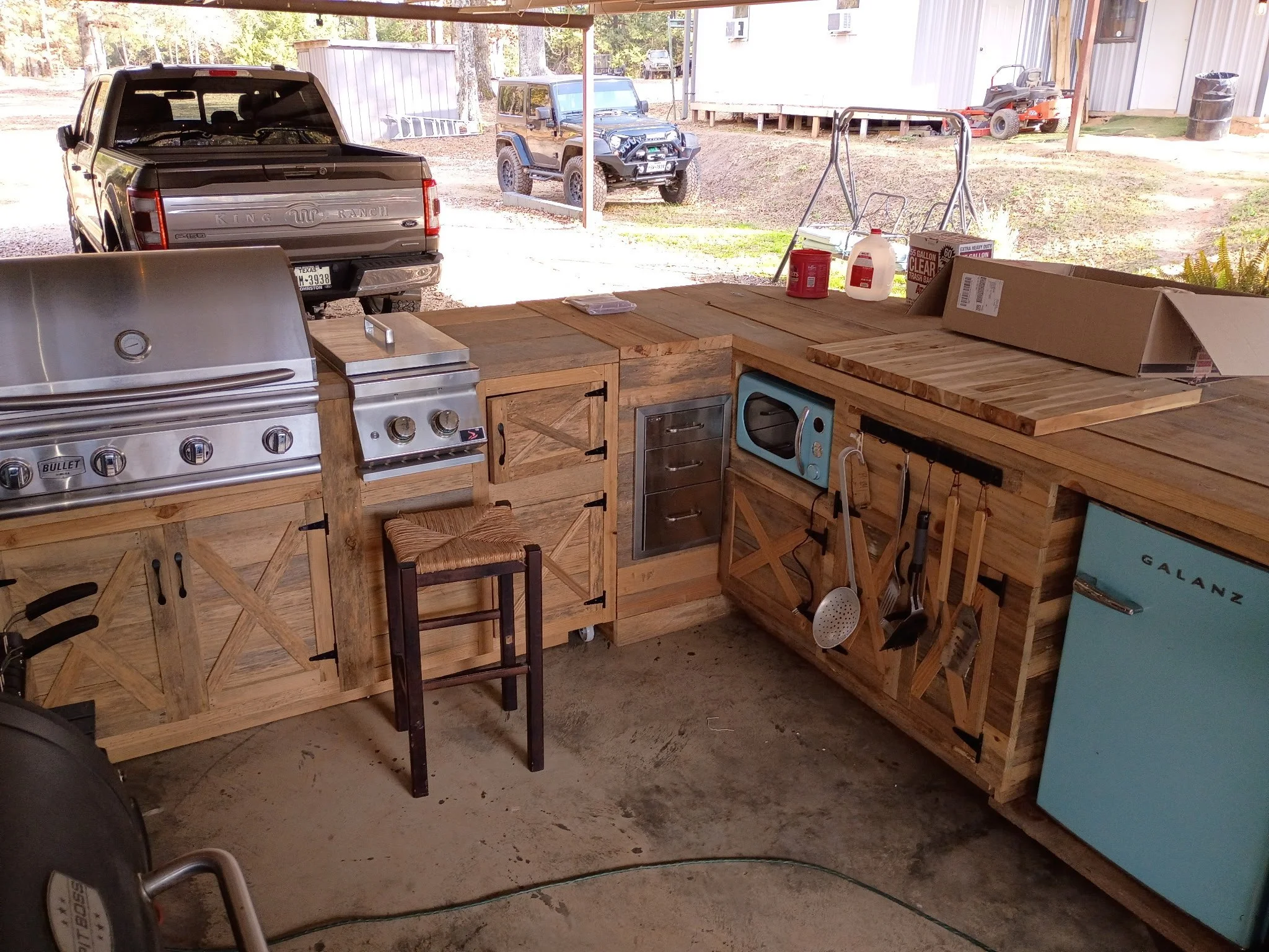 Outdoor kitchen with wood cabinets and stainless steel appliances, including a grill, stove, microwave, and small refrigerator, with vehicles and a shed visible in the background.