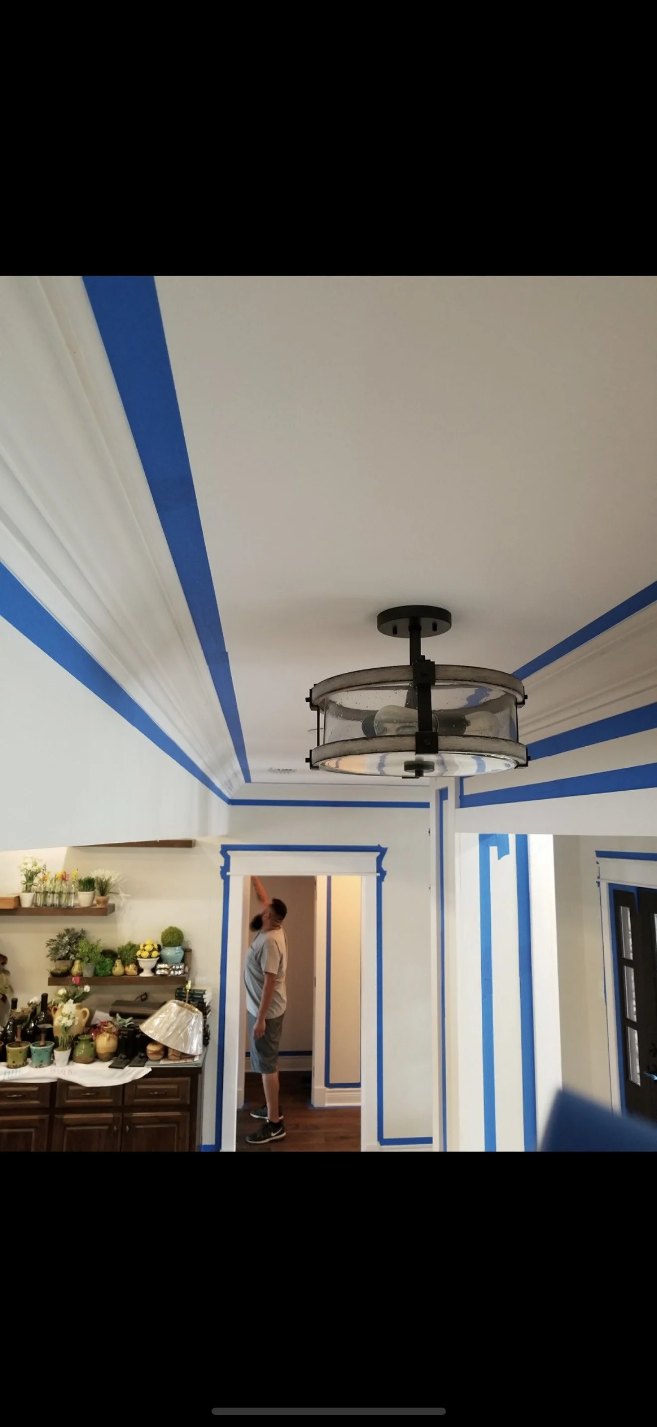 A room with white walls and blue painter's tape outlining the ceiling and door frames. There is a ceiling light fixture and a young boy wearing a gray t-shirt and shorts standing near a doorway, looking away. A counter with various decorative items a