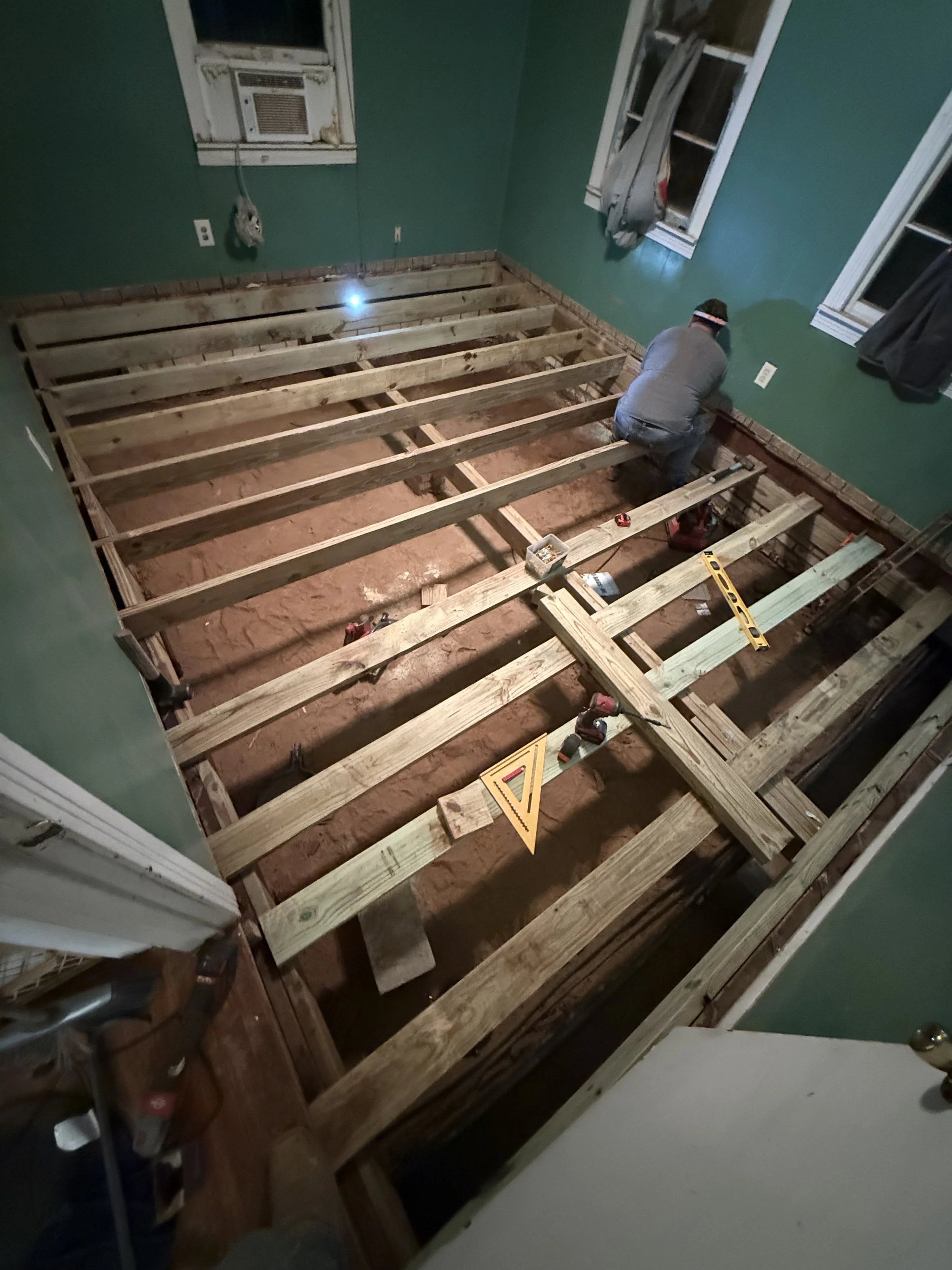 A person is working on the wooden framework of a floor in a room under renovation. The room has green walls, two windows with curtains, and an air conditioning unit. Tools and a spirit level are visible on the wooden beams.