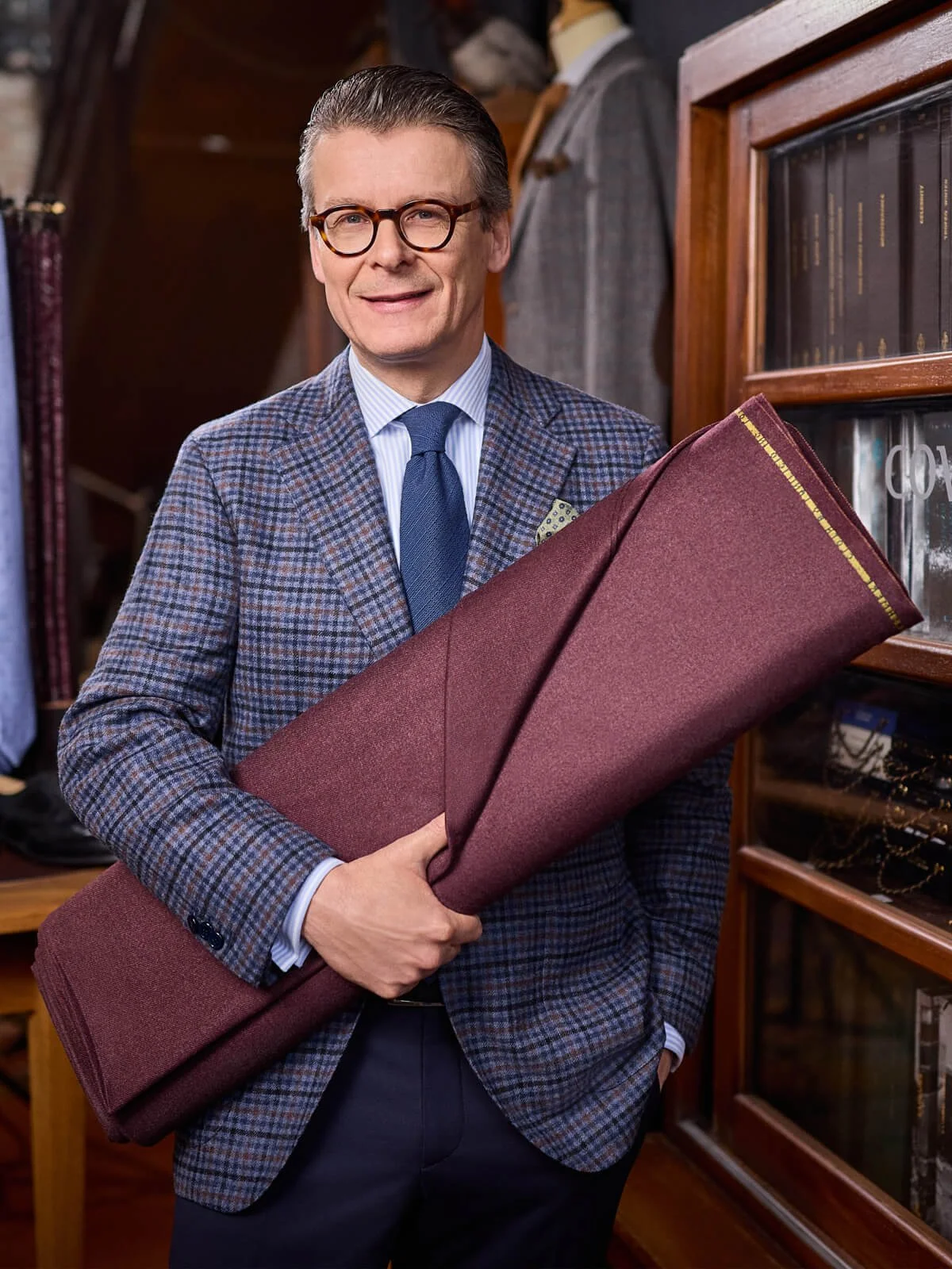 Ein stilvoll gekleideter Mann mit Brille hält ein großes rotes Fensterrollo in der Hand. Im Hintergrund sind Bücherregale und Anzüge zu sehen.