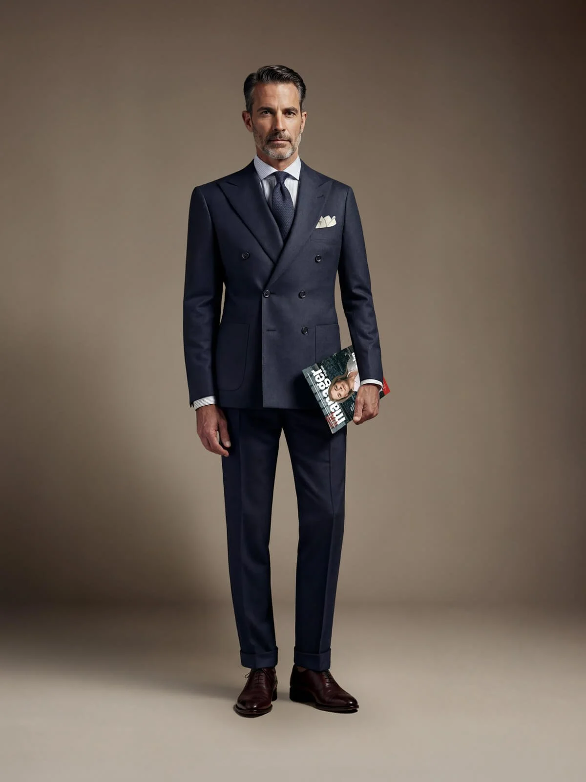 Ein Mann in einem dunkelblauen, zweireihigen Anzug steht vor einem neutralen Hintergrund, hält eine Zeitschrift in der linken Hand und schaut ernst in die Kamera.