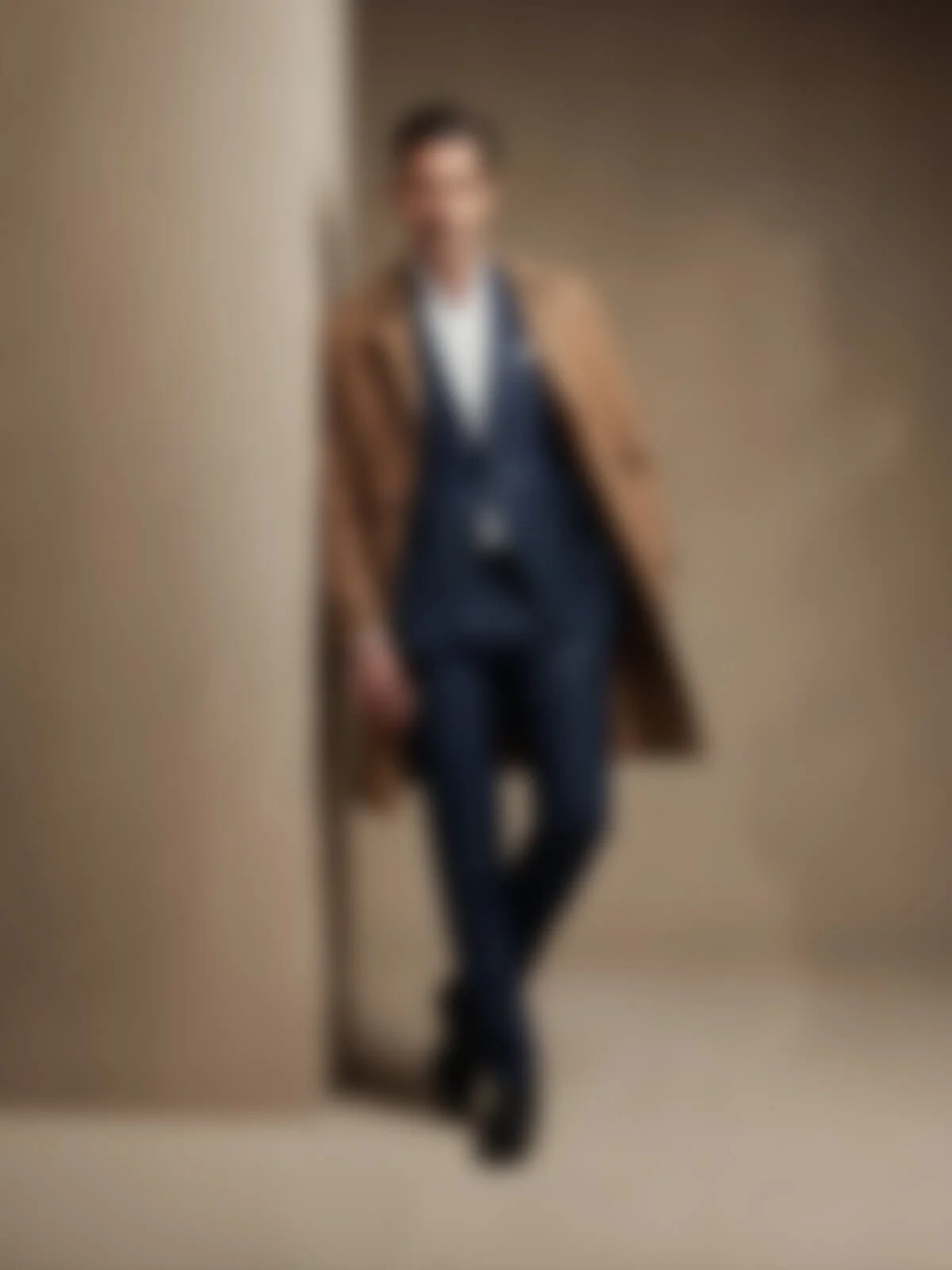 Ein Mann trägt einen braunen Mantel, eine dunkle Anzughose, eine weiße Hemd und eine schwarze Krawatte, lehnt an einer Wand in einem hellen Raum.