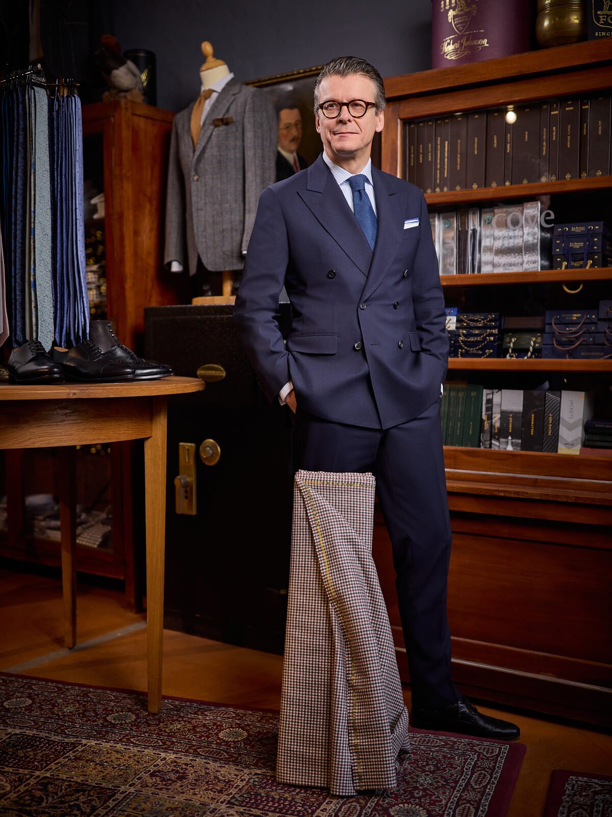 Ein stilvoll gekleideter Mann steht in einem Herrengeschäft, umgeben von Anzügen, Schuhen und Accessoires, mit einem leichten Lächeln.