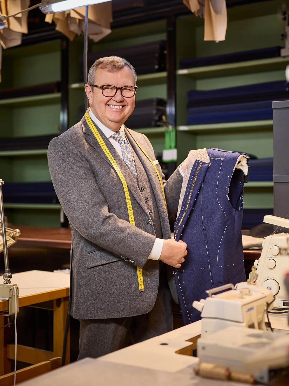 Ein Mann trägt einen grauen Anzug und eine Brille, hält ein Schneiderpult mit einem angefertigten blauen Anzug und lächelt in der Schneiderei."}