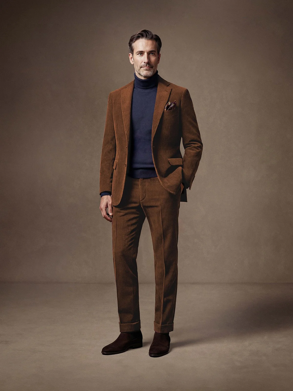 Ein Mann trägt einen braunen Anzug mit passender Hose, ein dunkelblauer Rollkragenpullover, braune Stiefel und hat eine stilvolle Haltung vor einem neutralen Hintergrund.