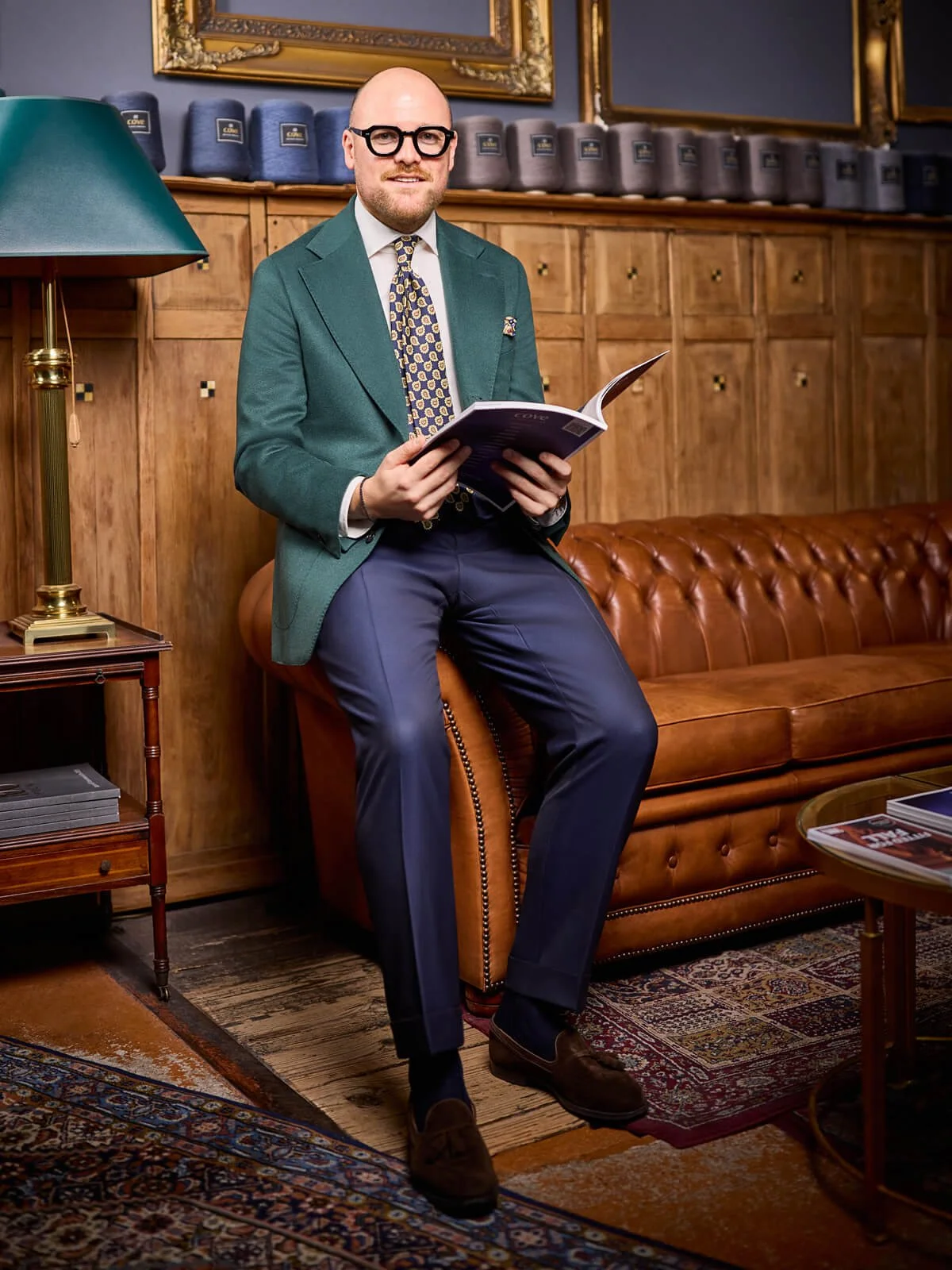 Ein Mann mit Brille, Anzug und Krawatte sitzt auf einem braunen Ledersofa in einem stilvollen, altmodischen Raum und liest eine Zeitschrift.