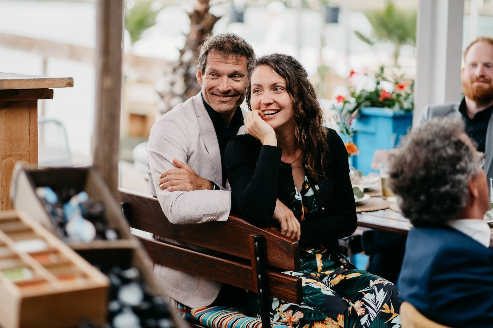 People sitting at a table outdoors at a social gathering or event, smiling and engaging with each other.