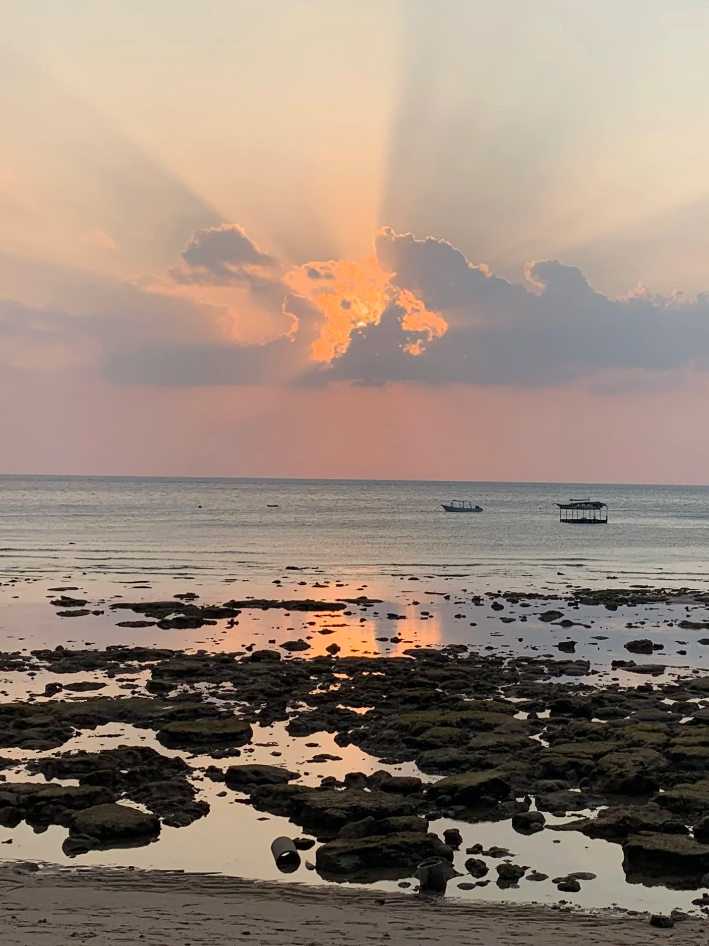 Sunset over the ocean with clouds partially blocking the sun, creating rays of light. Rocky shoreline in the foreground with water reflections, and boats floating on the water.