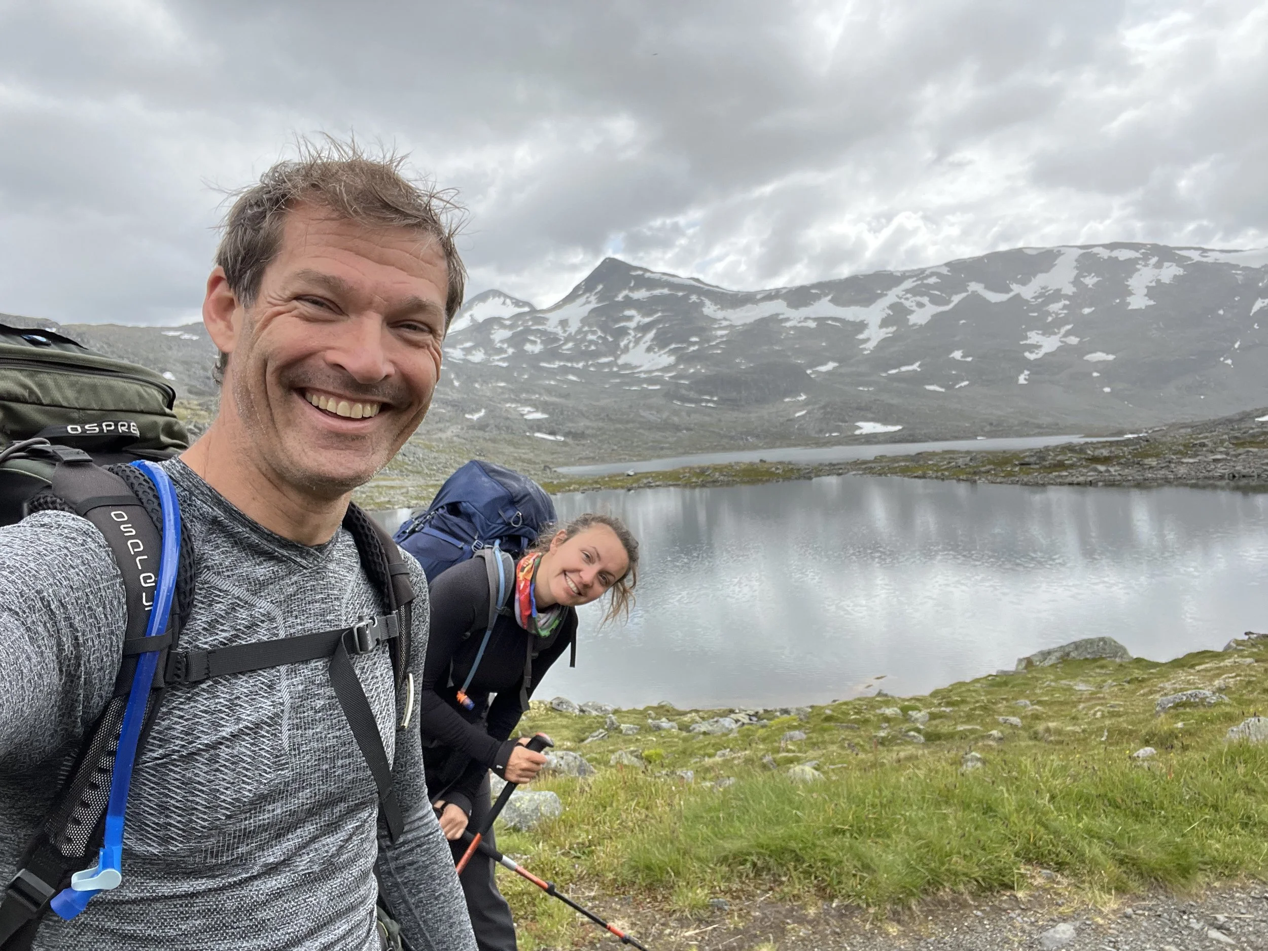 Two hikers, a man and a woman, smiling with backpacks and trekking poles beside a lake in a mountainous area with snow patches and cloudy sky.